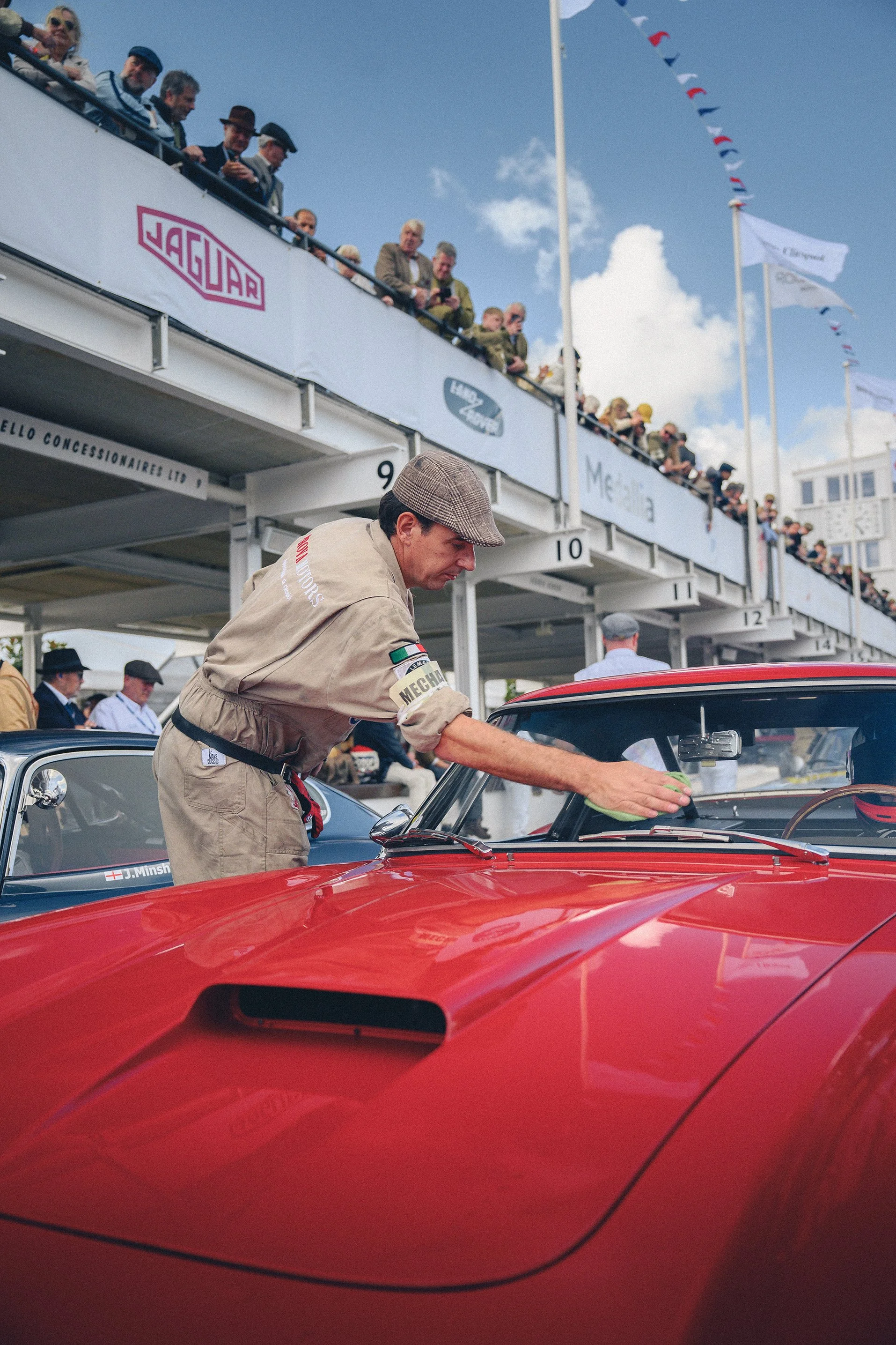 A man in vintage racing attire is cleaning a red classic car at a car show or race event, with an audience watching from a balcony above under a partly cloudy sky.