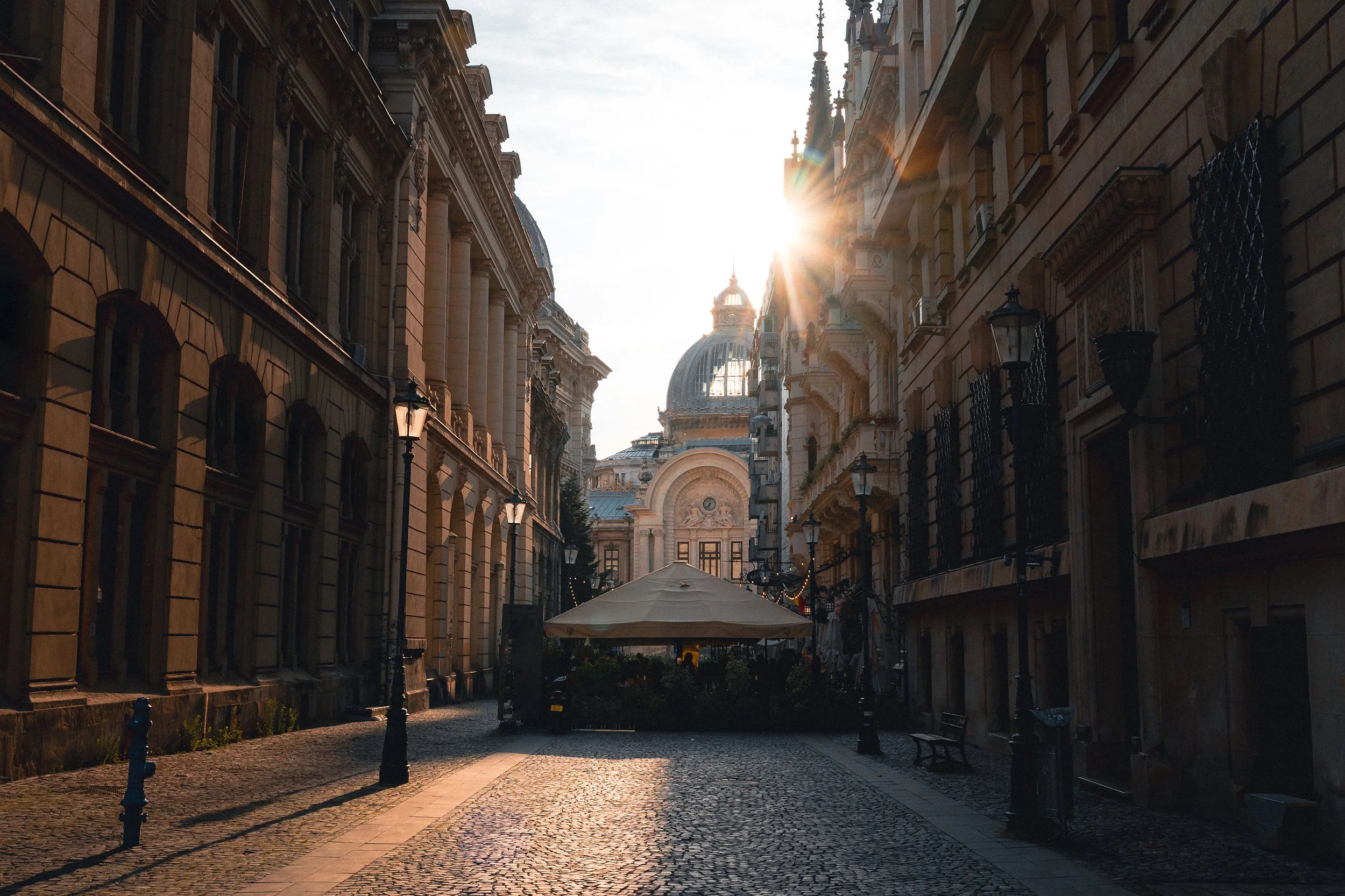A cobblestone street in a historic European city, bathed in warm sunlight with old buildings on both sides, and a domed building in the background.
