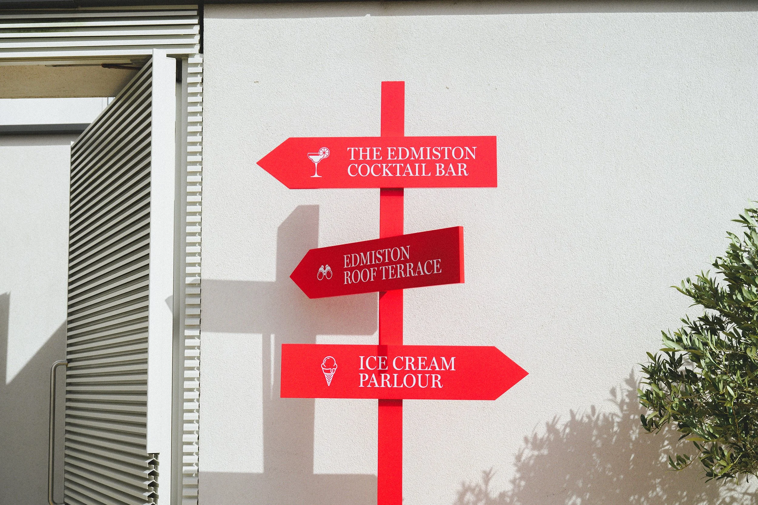 Red directional signpost with three signs pointing to the left and right, indicating locations such as The Edmiston Cocktail Bar, Edmiston Roof Terrace, and Ice Cream Parlour, mounted on a white wall with shadows, next to some greenery.
