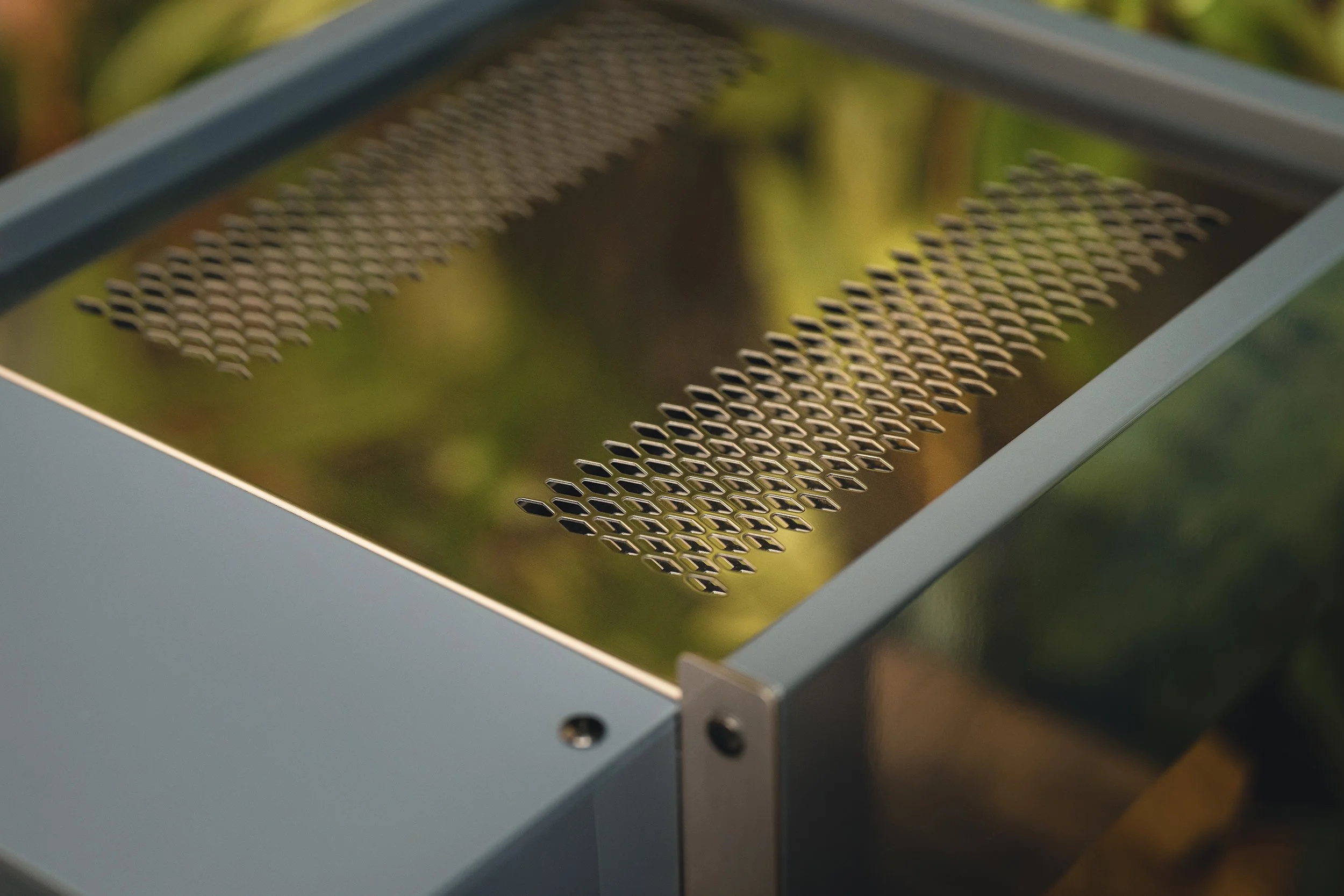 Close-up of a metal grater with perforated holes, set on a reflective surface with blurred green foliage in the background.