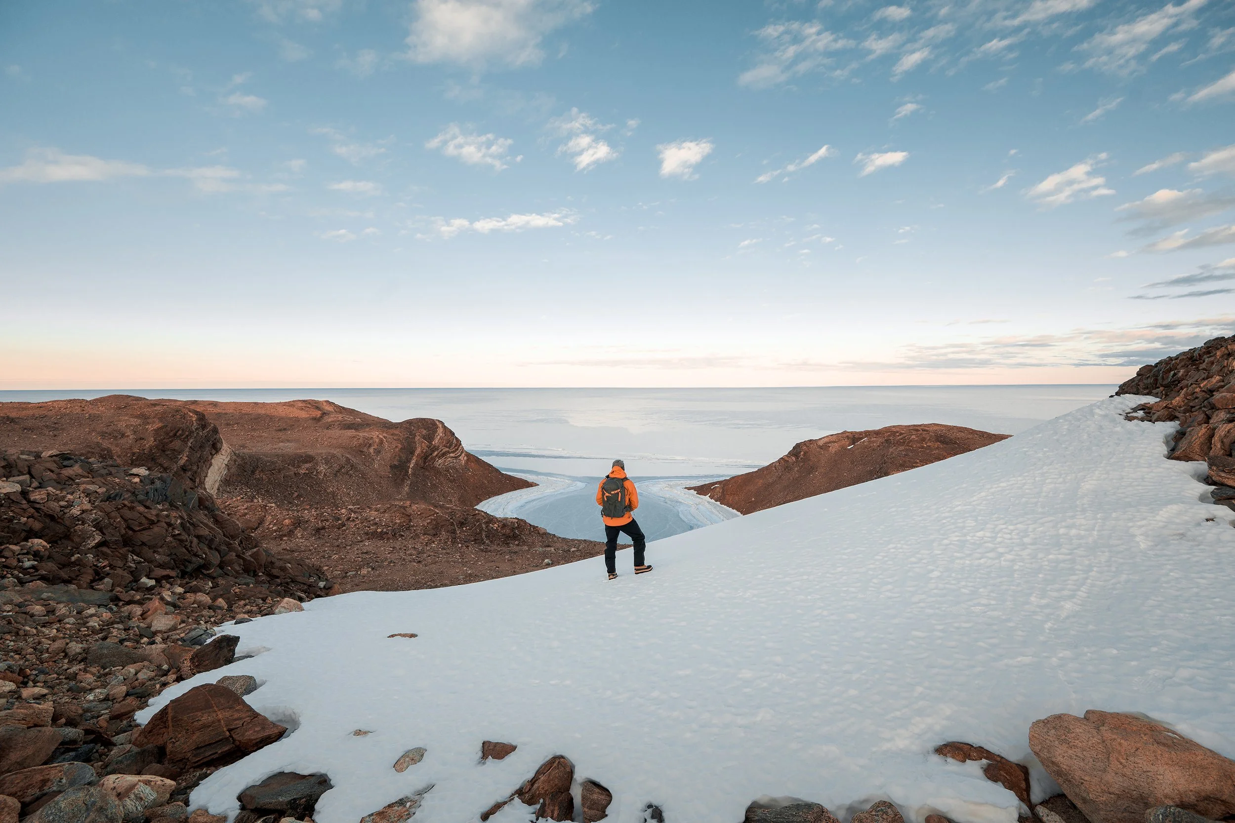 A hiker in an orange jacket and backpack walking on snow-covered terrain between rocky hills towards a flat icy landscape under a partly cloudy sky.