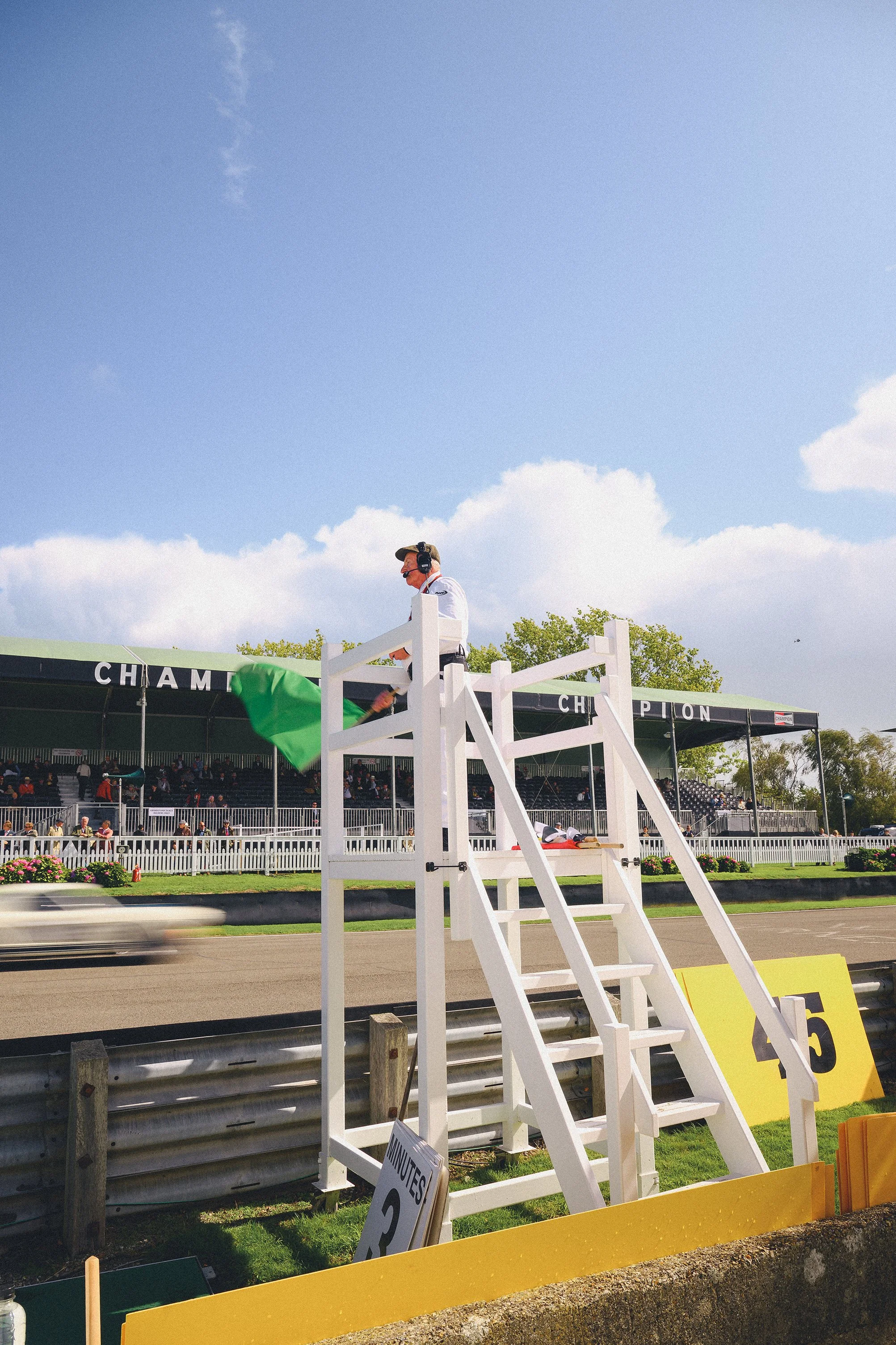 A male race official holding a green flag at a horse racing track, standing on a white platform near the finish line with a crowd and racecourse visible in the background.