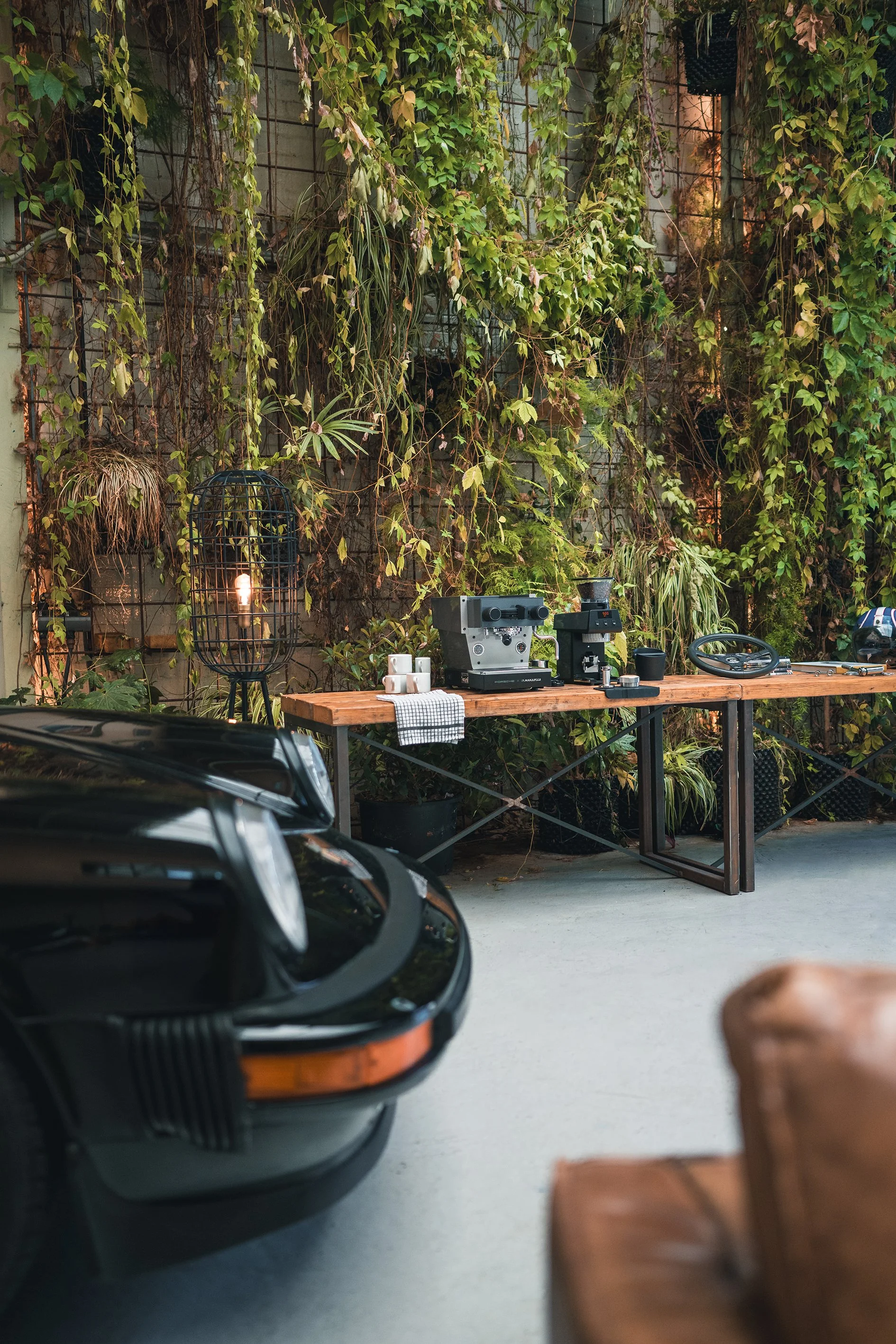 A black vintage car in the foreground with part of a brown leather chair visible on the right side, and a wooden table on the back wall holding a coffee machine, a grinder, cups, and a lamp with greenery in the background.