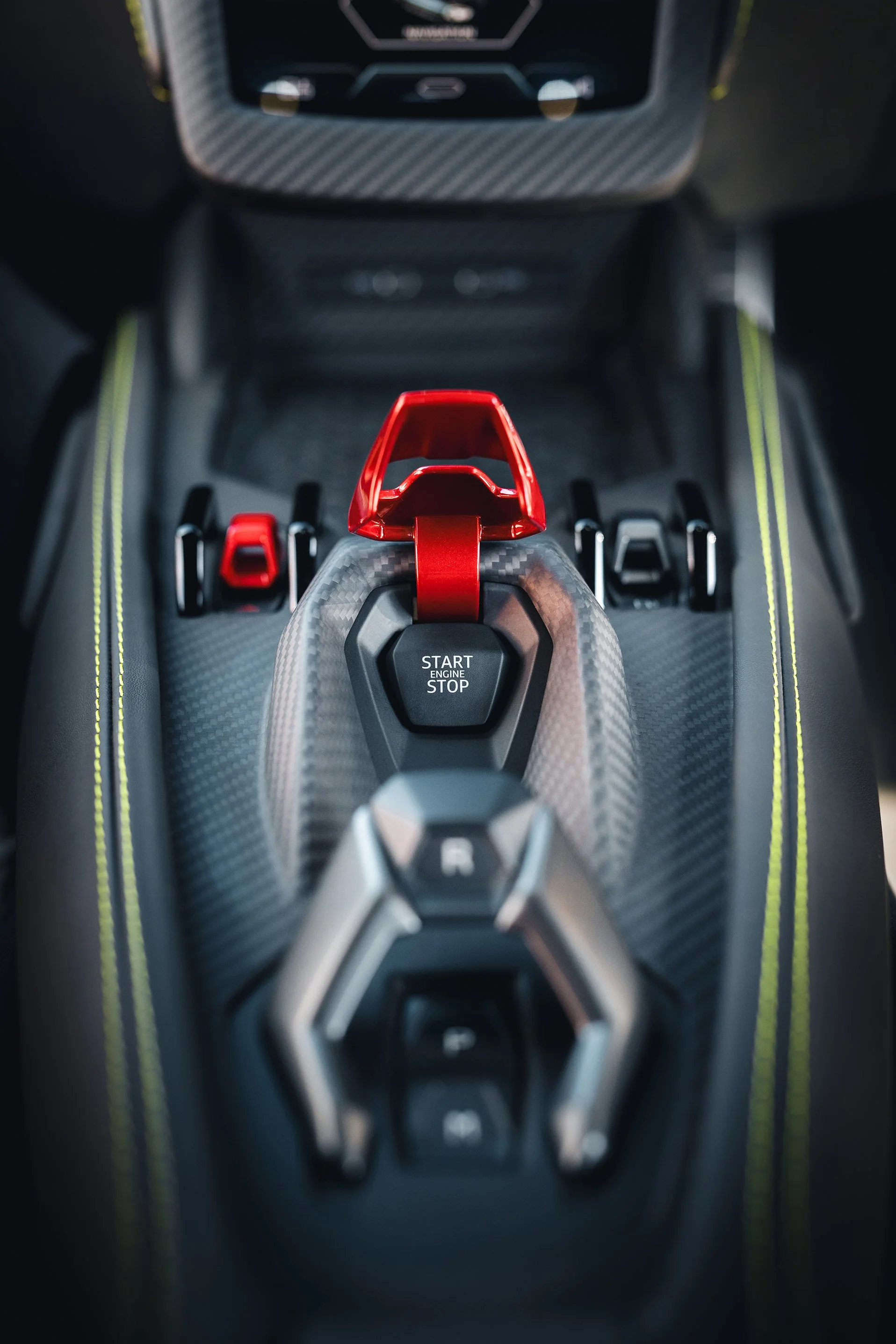 Close-up of the center console of a sports car, featuring a red gear shift lever, a start/stop engine button, and various control switches, with carbon fiber trim and accented stitching.