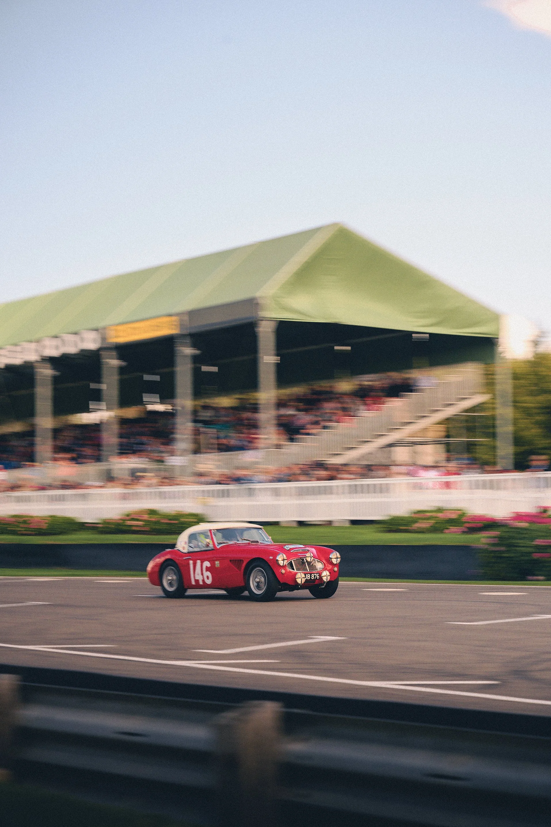 A red vintage race car with number 146 on the side speeding on a racetrack, with a green grandstand filled with spectators in the background.