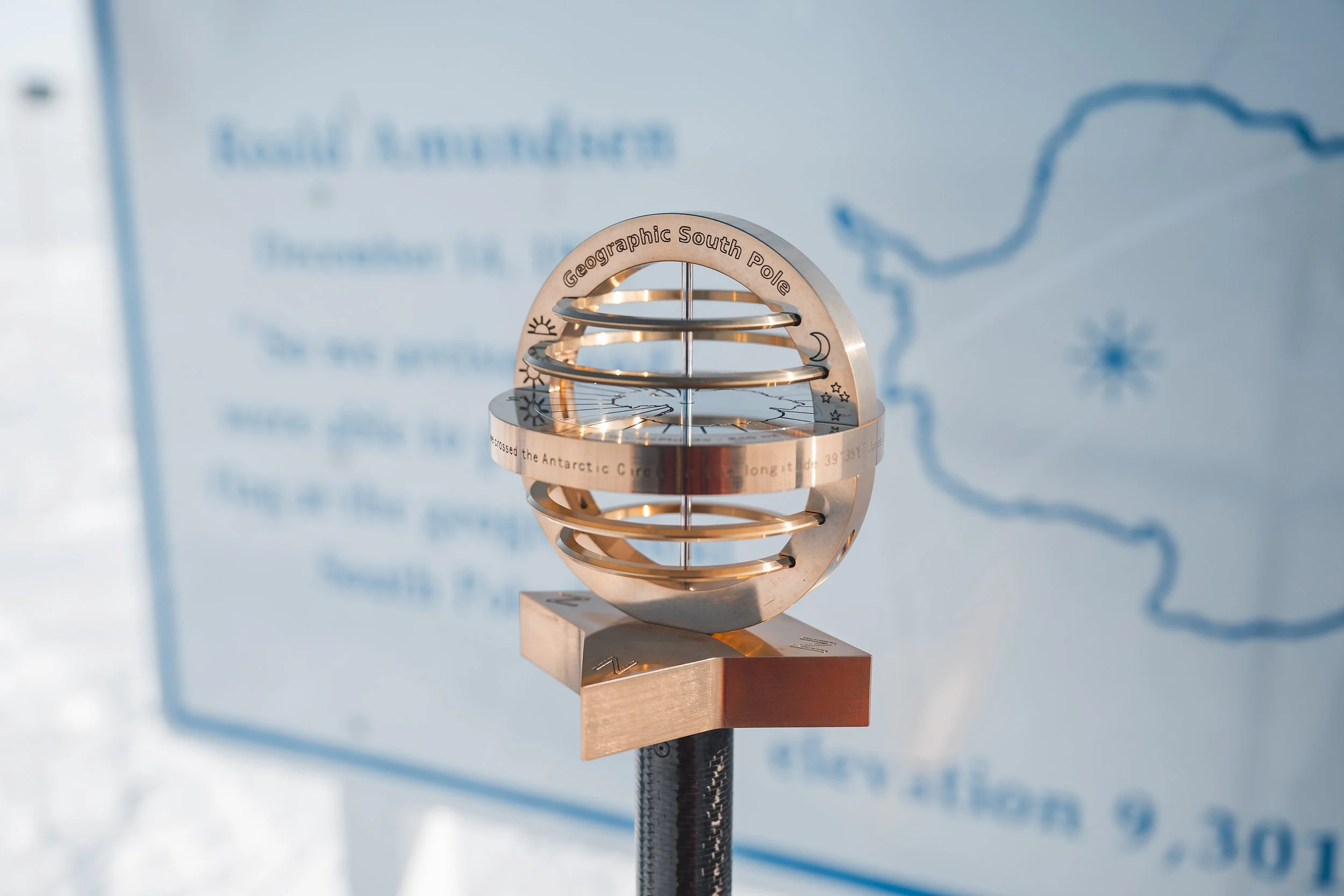 A metallic globe-shaped marker on a black pole, labeled 'Geographic South Pole,' with a background of a blue map.
