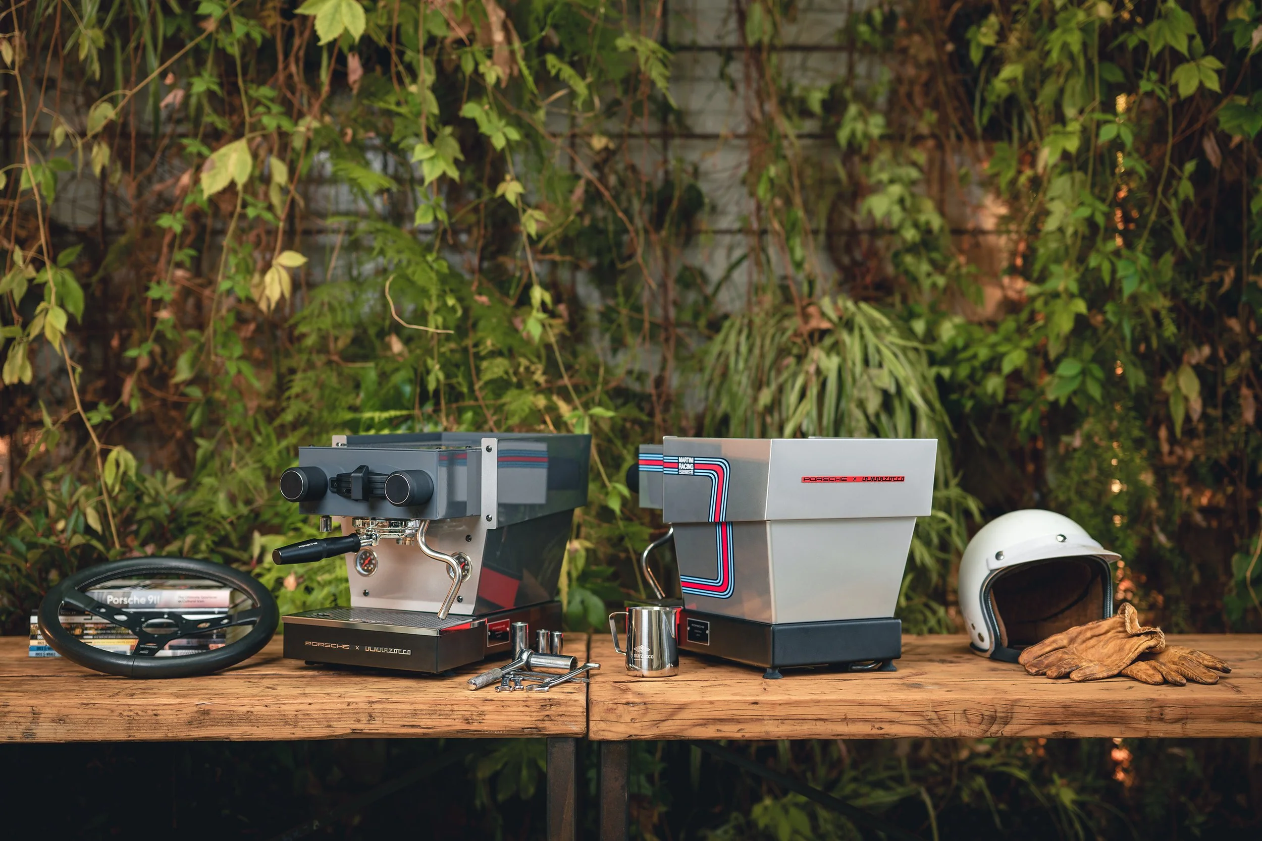 A wooden table outdoors with two espresso machines, a racing helmet, gloves, and a small metal pitcher, set against a background of green foliage.