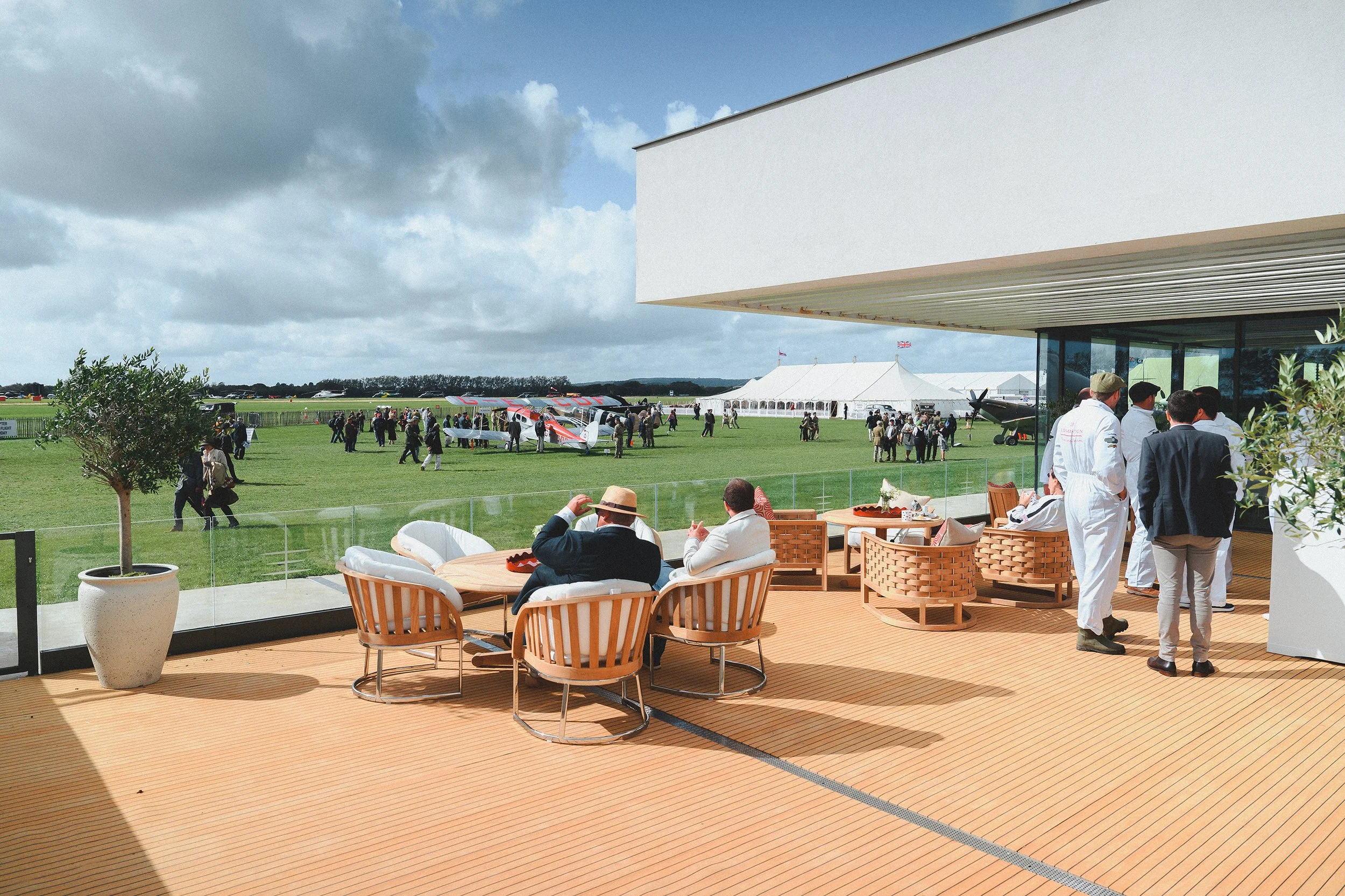 Lounge area overlooking an airfield with vintage aircraft and a crowd outside a large tent, under partly cloudy skies.