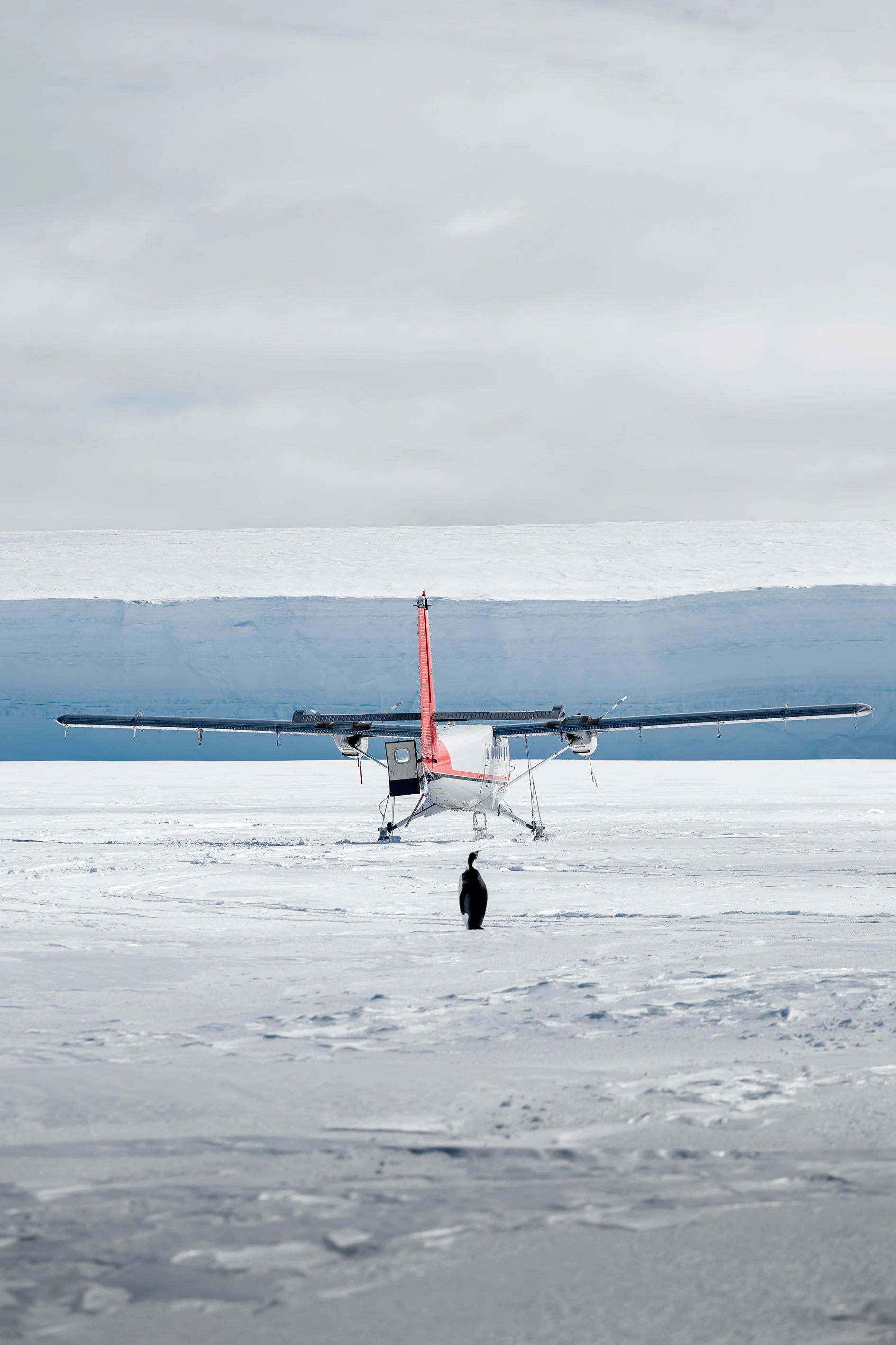 A small aircraft on snow in Antarctica with a penguin in the foreground and ice-covered mountains in the background.