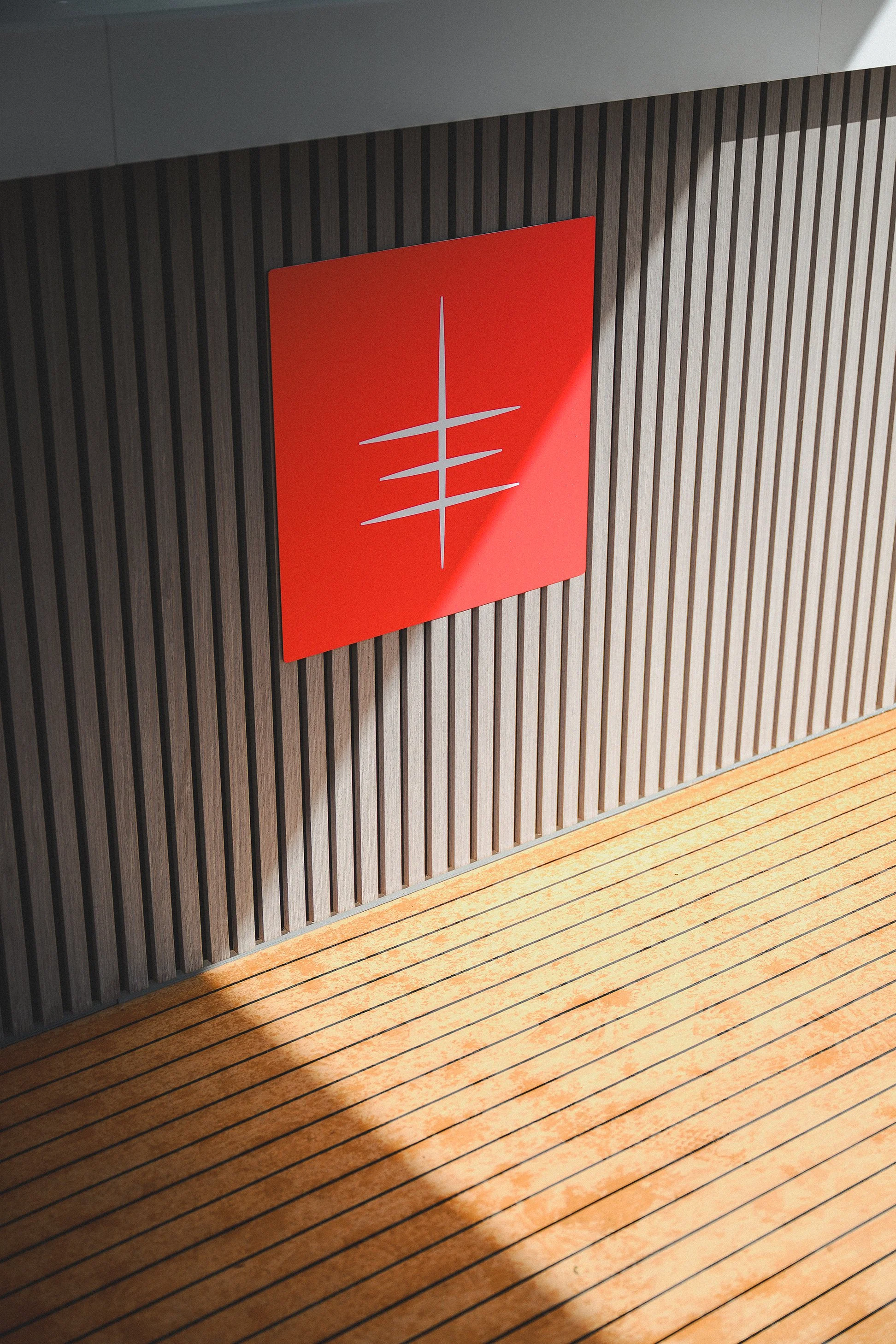 Red wall-mounted sign with a white abstract logo. The sign is on a wall with vertical wooden slats, near a wooden surface with horizontal stripes.