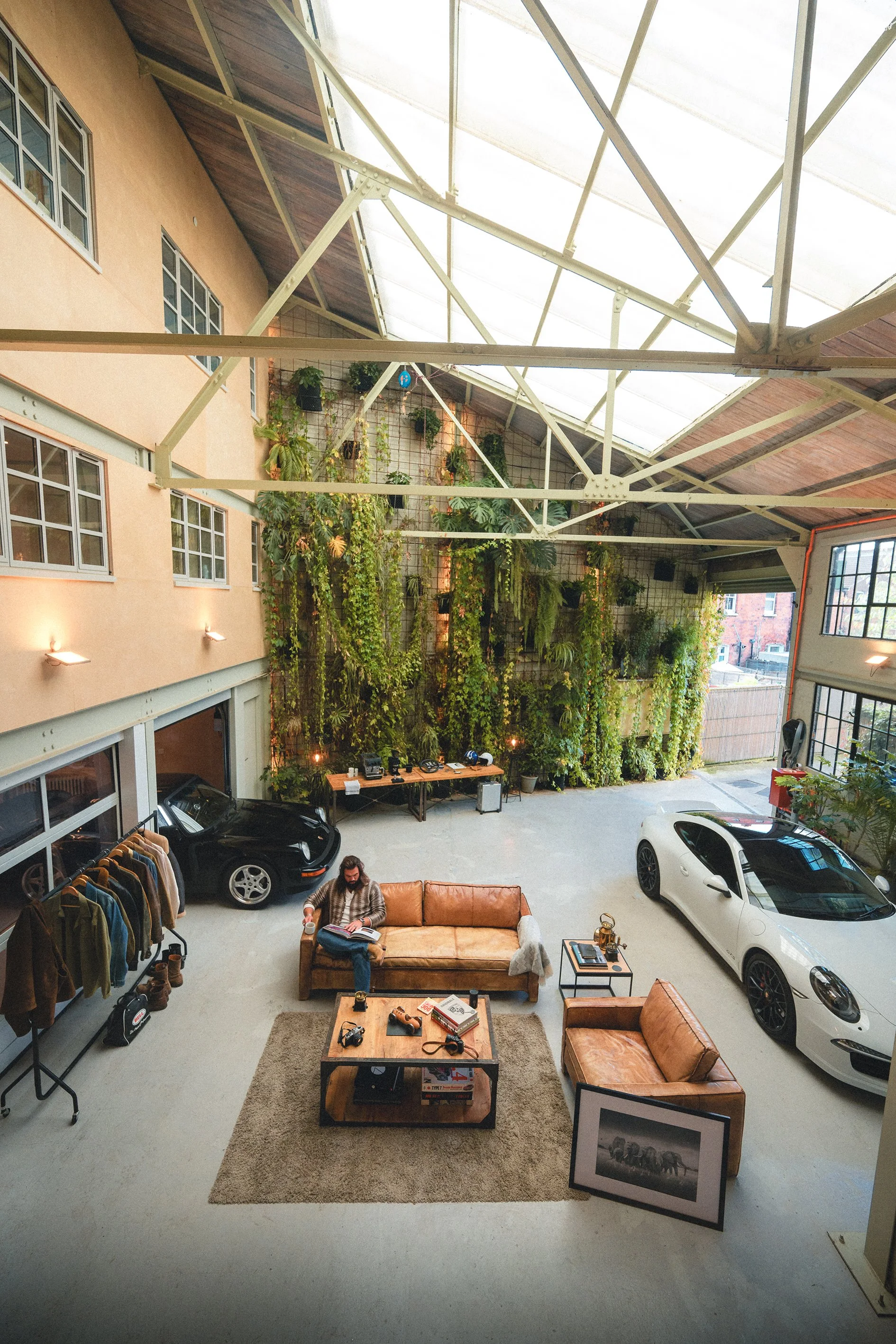 Interior of a spacious garage converted into a modern lounge with vintage furniture, cars, and a green wall of plants.