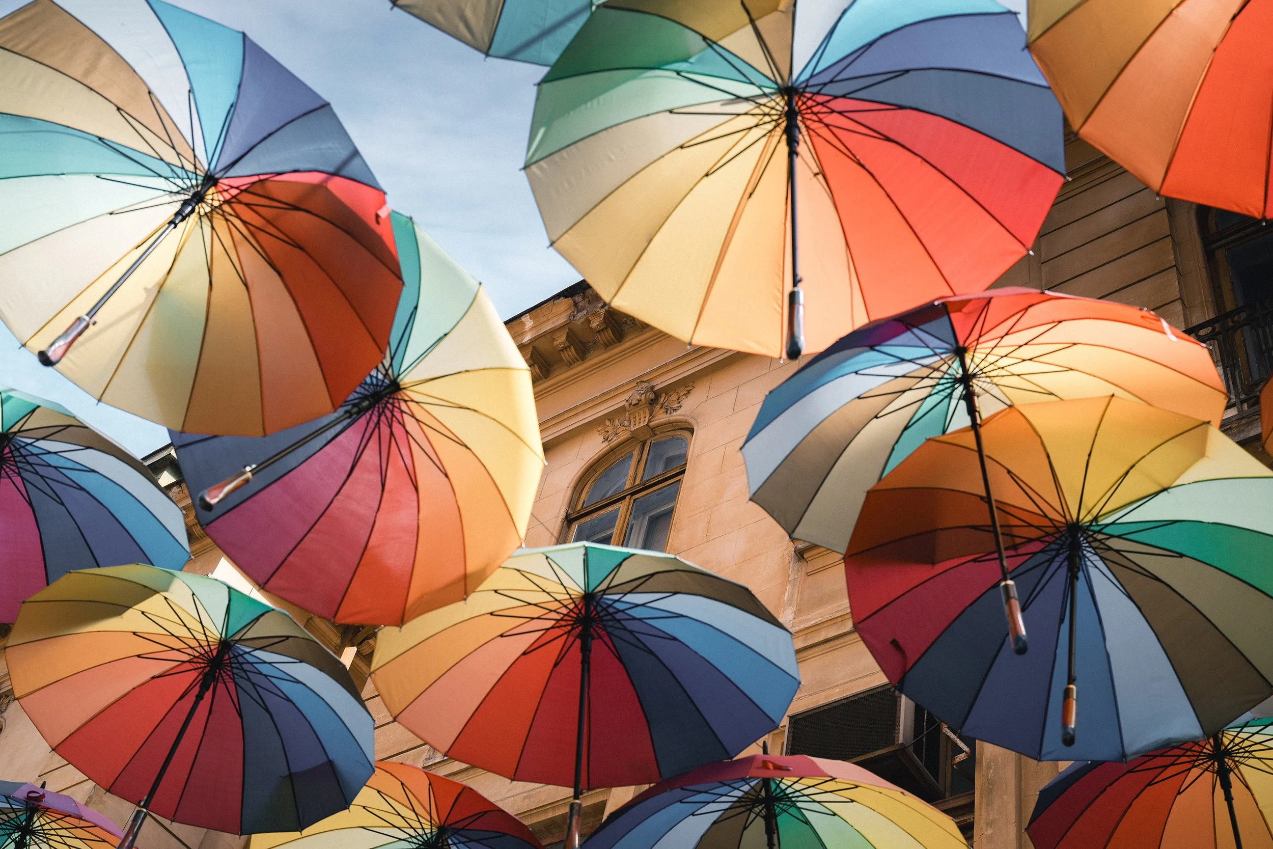 Colorful umbrellas suspended upside down outdoors in front of a building with ornate windows.
