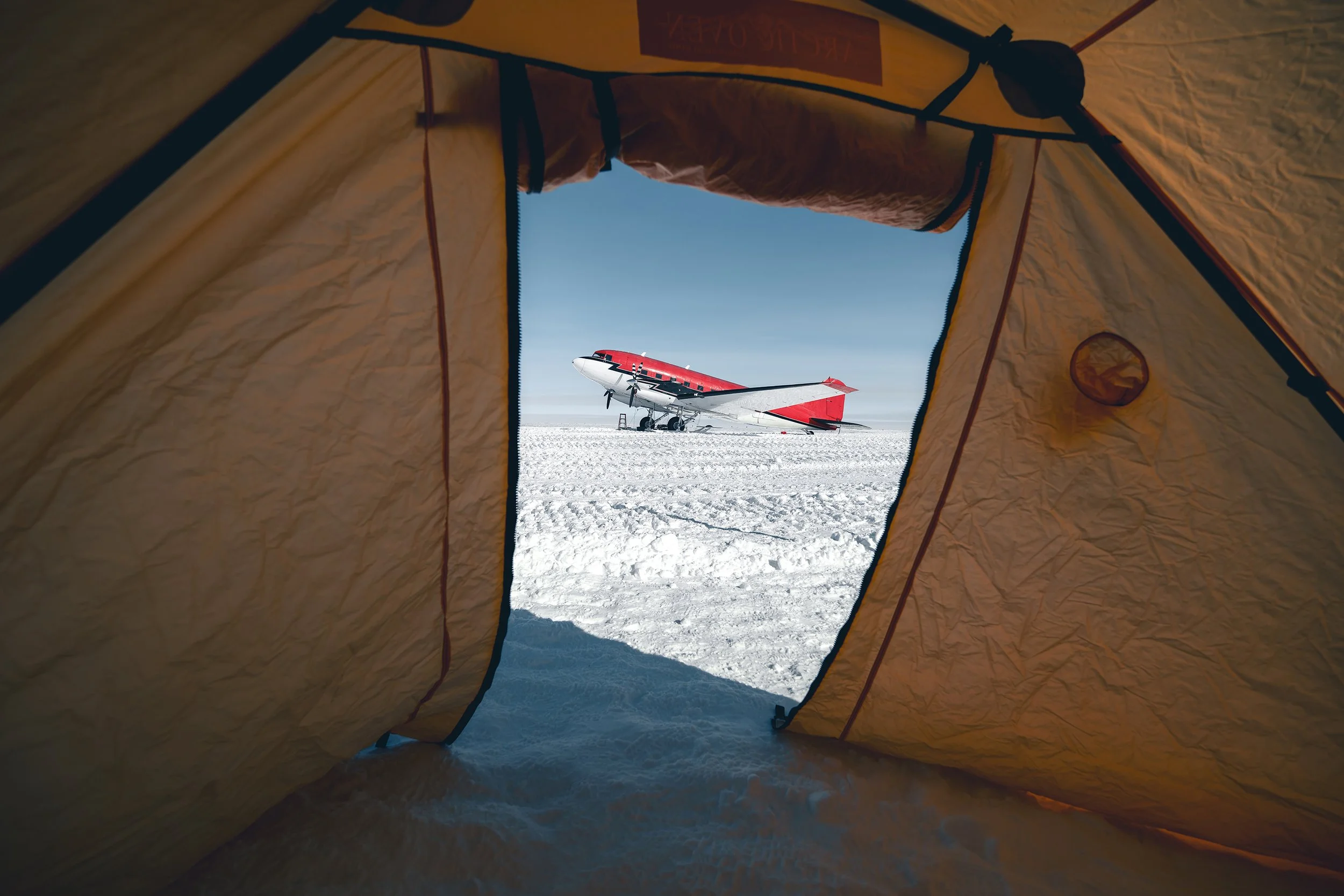 Looking out from inside a camp tent onto an airplane on a snow-covered landscape under a clear blue sky.