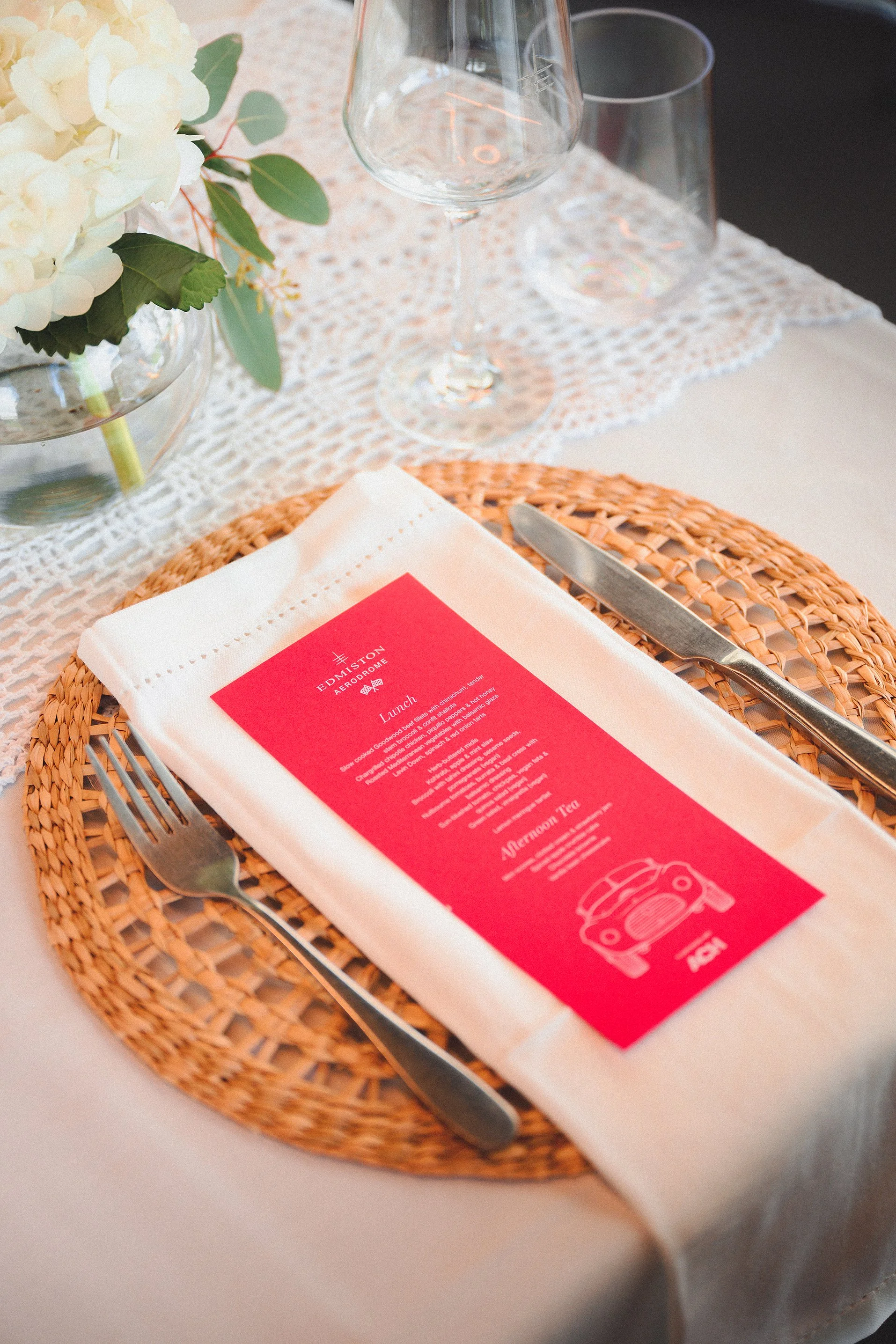 A place setting on a dining table with a woven placemat, white napkin, stainless steel fork and knife, a menu with a red cover, a glass of water, a wine glass, and a floral centerpiece.