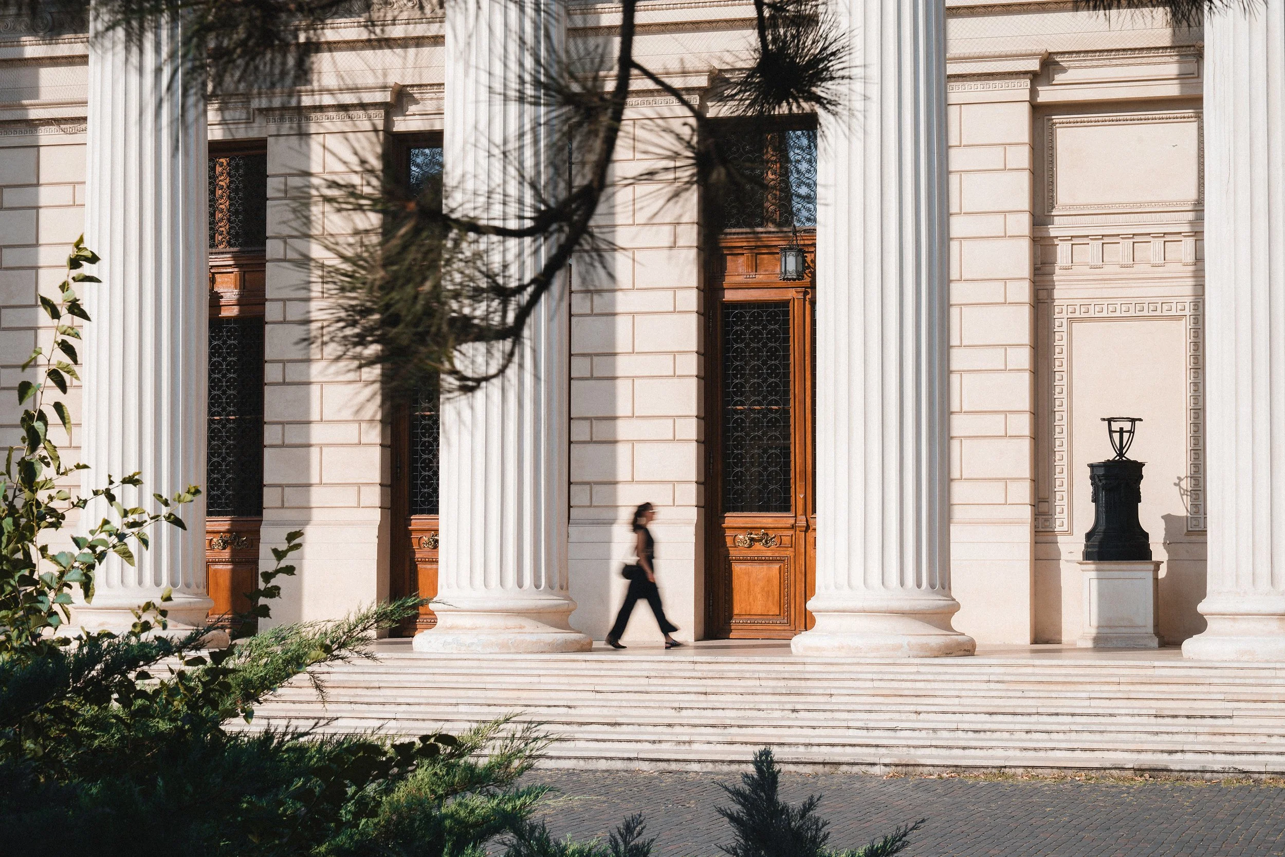 A person in black walking past the grand entrance of a neoclassical building with large white columns, wooden doors, and steps leading up to the doorway.
