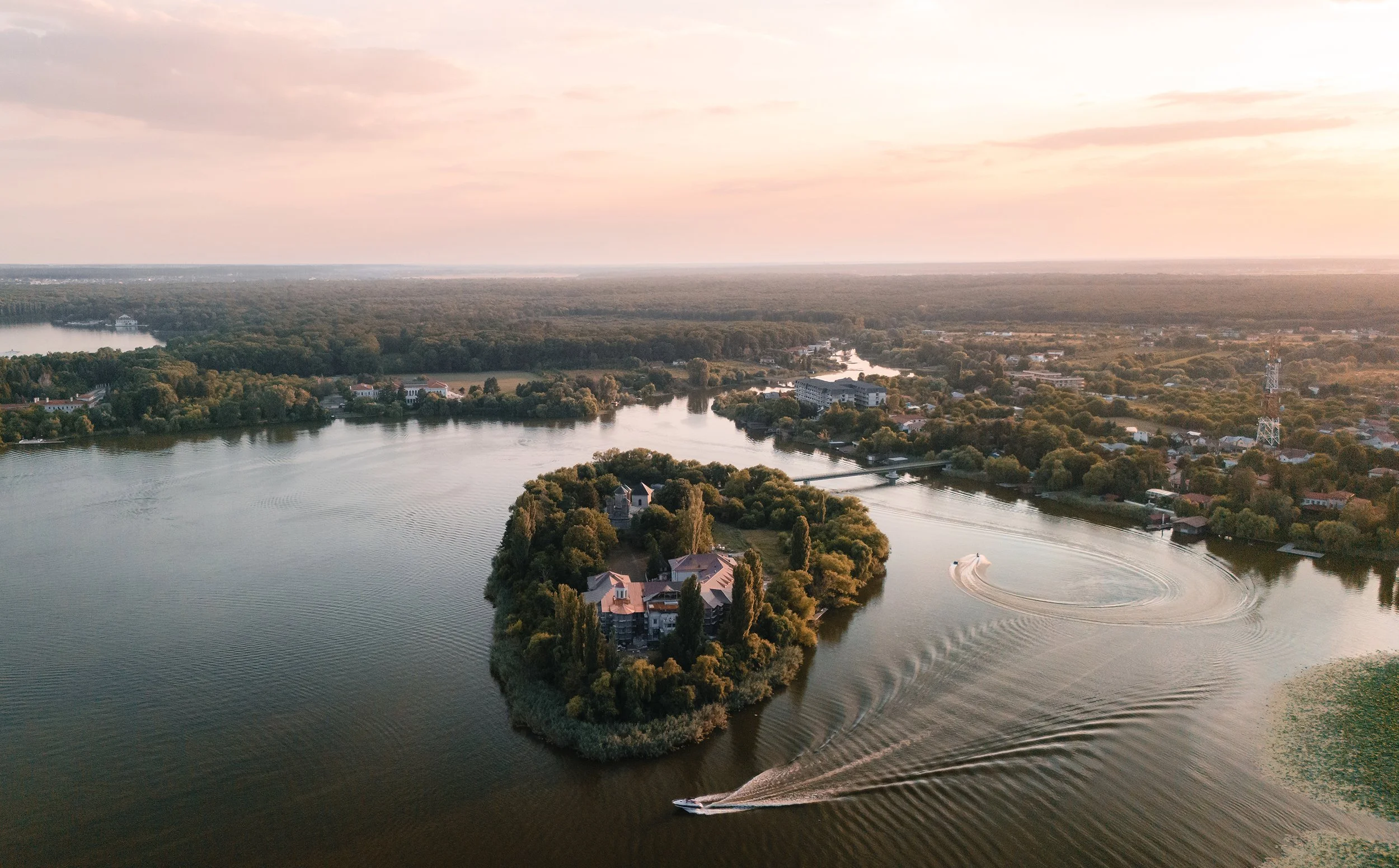 Aerial view of a river with an island featuring buildings and trees, a boat creating ripples in water, and a distant town with green vegetation at sunset.