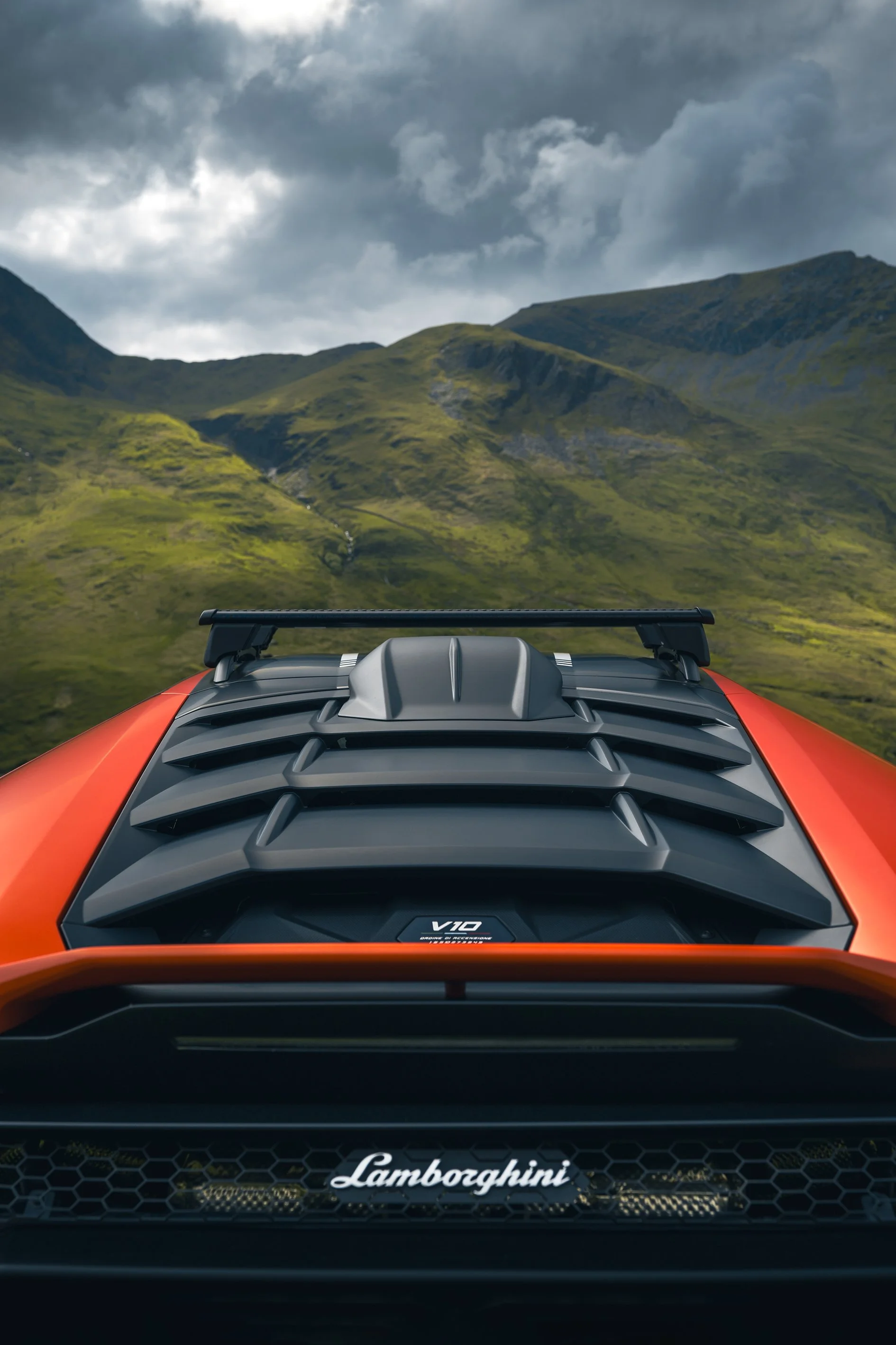 Front view of a Lamborghini supercar with an orange exterior, showing the black engine cover and the Lamborghini logo, with green mountains and cloudy sky in the background.