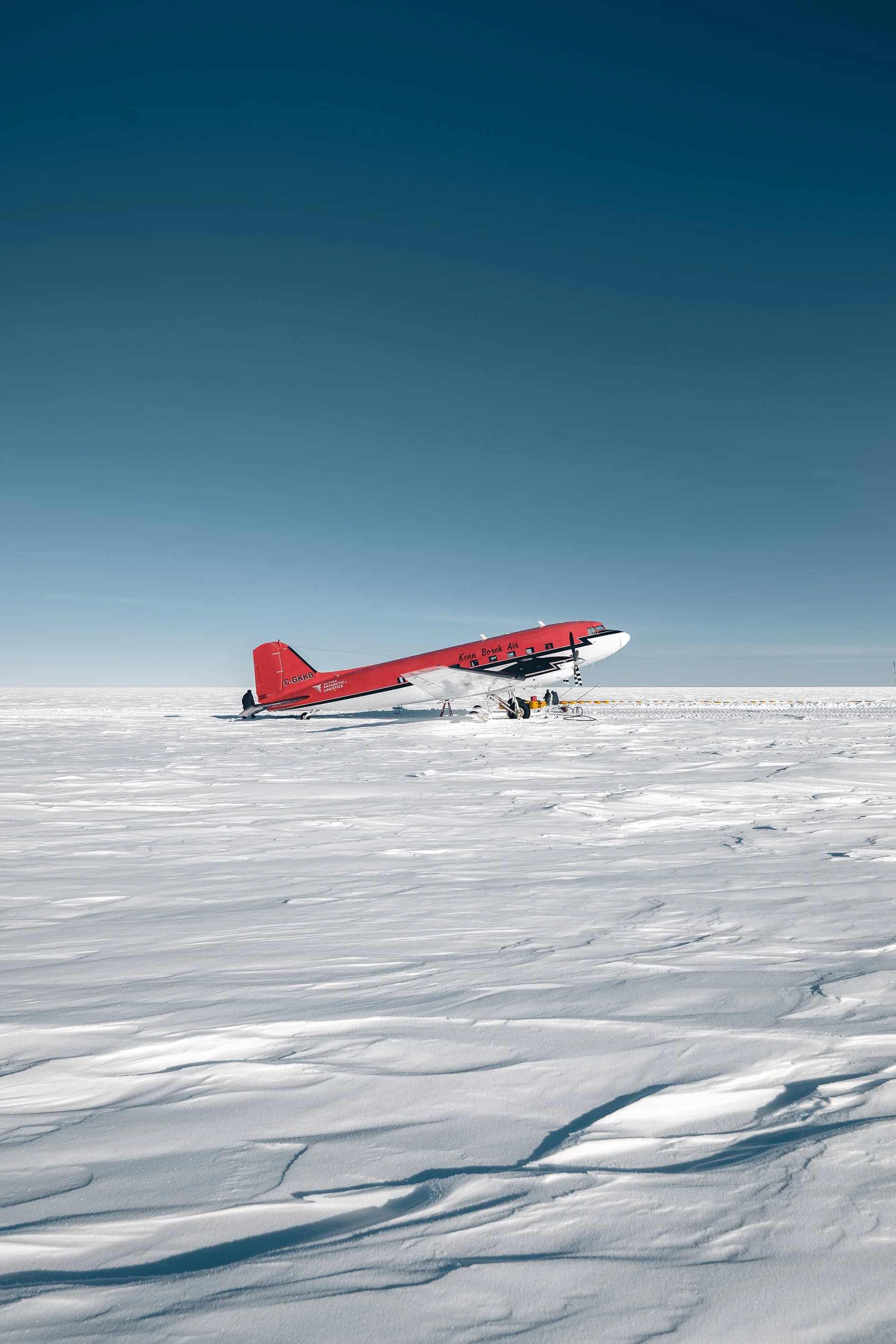 A red airplane on the snow-covered ground in a vast icy landscape under a clear blue sky.