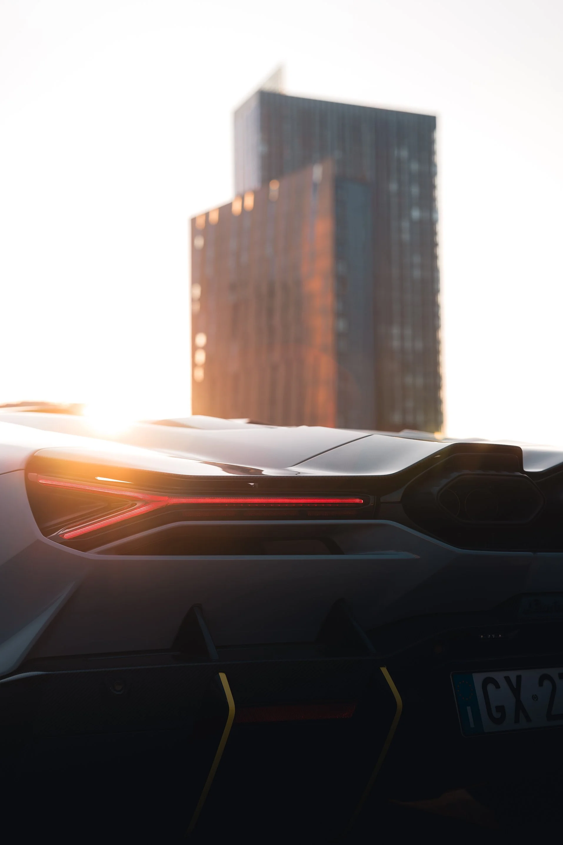 Close-up of a sleek sports car's rear with glowing taillights, with a modern high-rise building in the background during sunset.