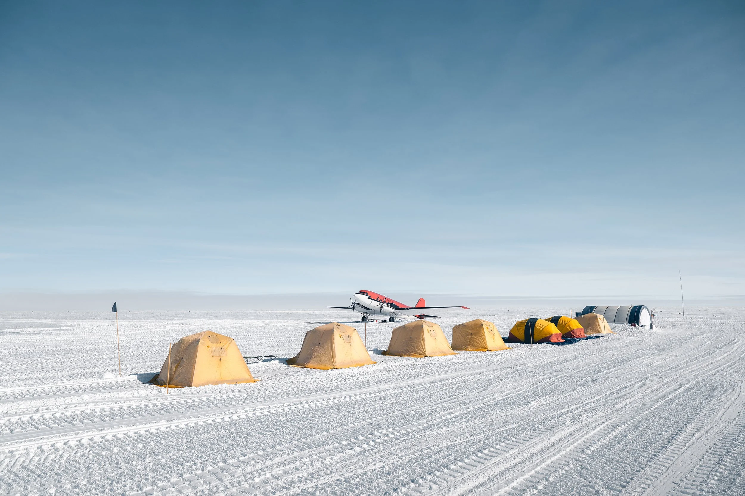 A snow-covered field with a row of yellow tents, a small aircraft in the background, and a curved hangar structure on the right, under a clear blue sky.