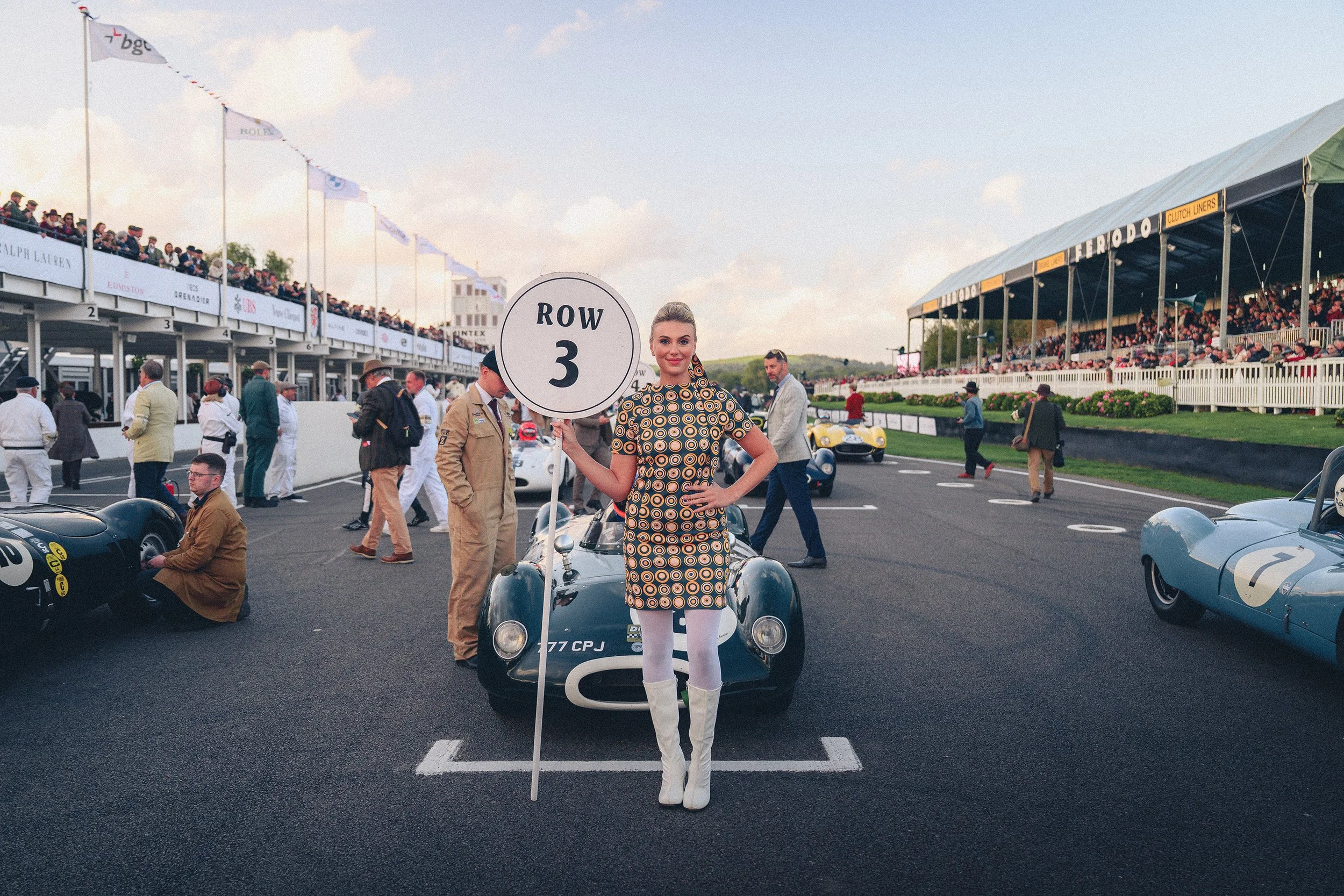 A woman holding a sign that reads 'ROW 3' stands in front of a vintage race car at a racing event. There are people, cars, and grandstands in the background.