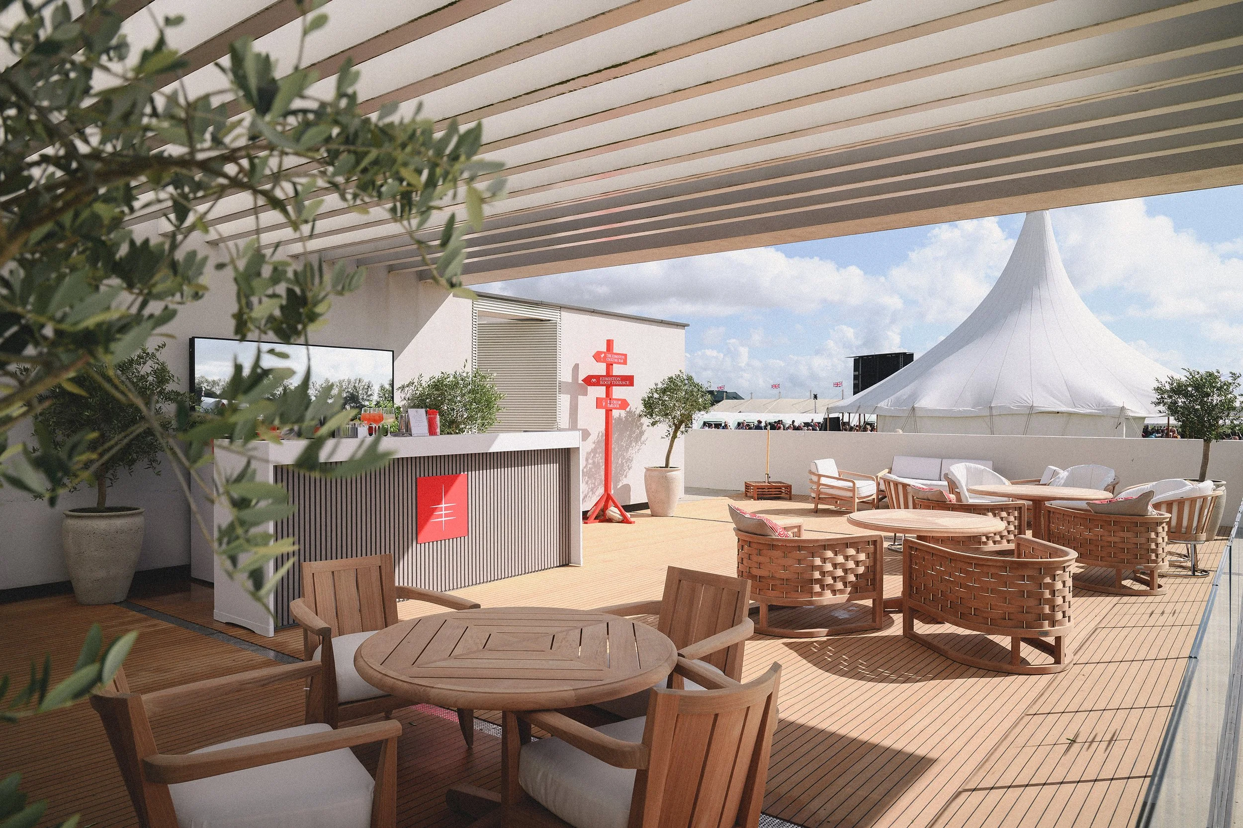 Outdoor seating area at a festival or event, with wooden chairs and tables, potted trees, a white tent, and a signpost with red arrows.
