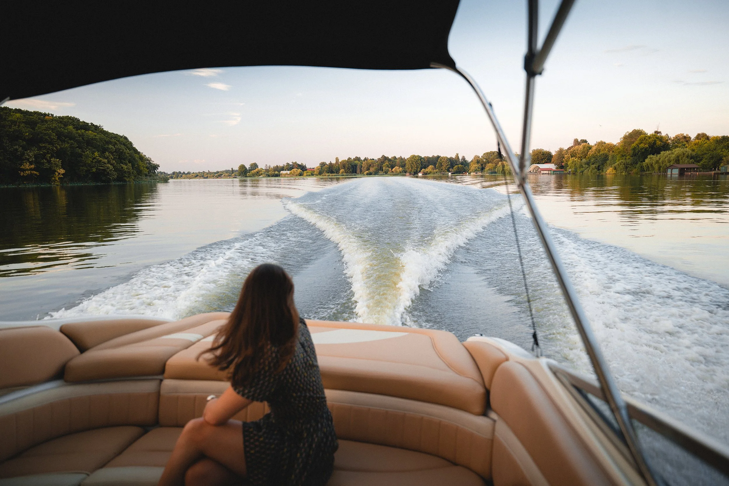 A woman sitting on a beige cushioned boat seat, looking out over the water while on a boat traveling along a river during sunset. The water is calm and reflects the trees and sky along the shoreline.