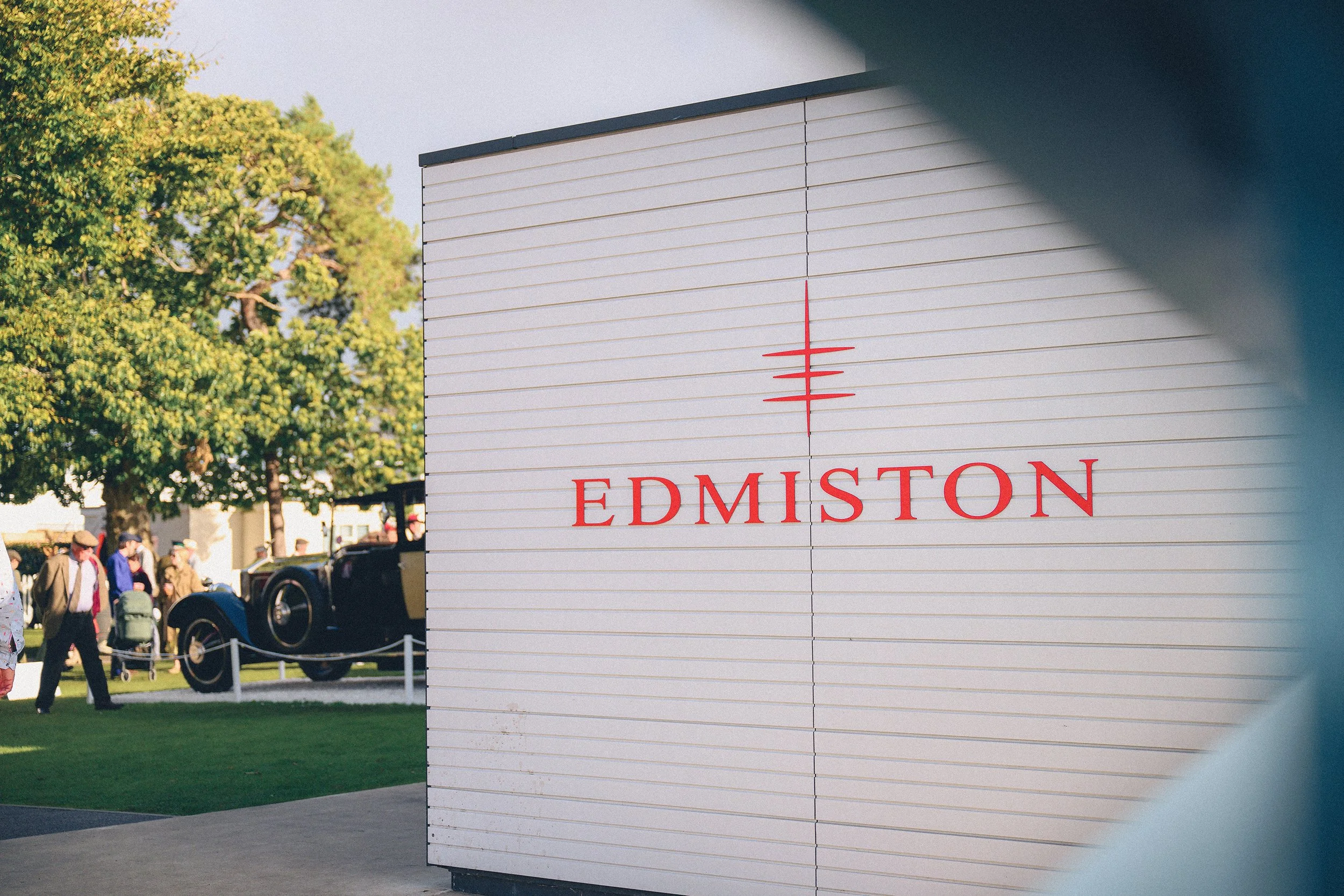 A large white sign with red text reading 'EDMISTON' and a simple red logo above it, positioned outdoors near a vintage car and people walking in a park-like setting with trees.