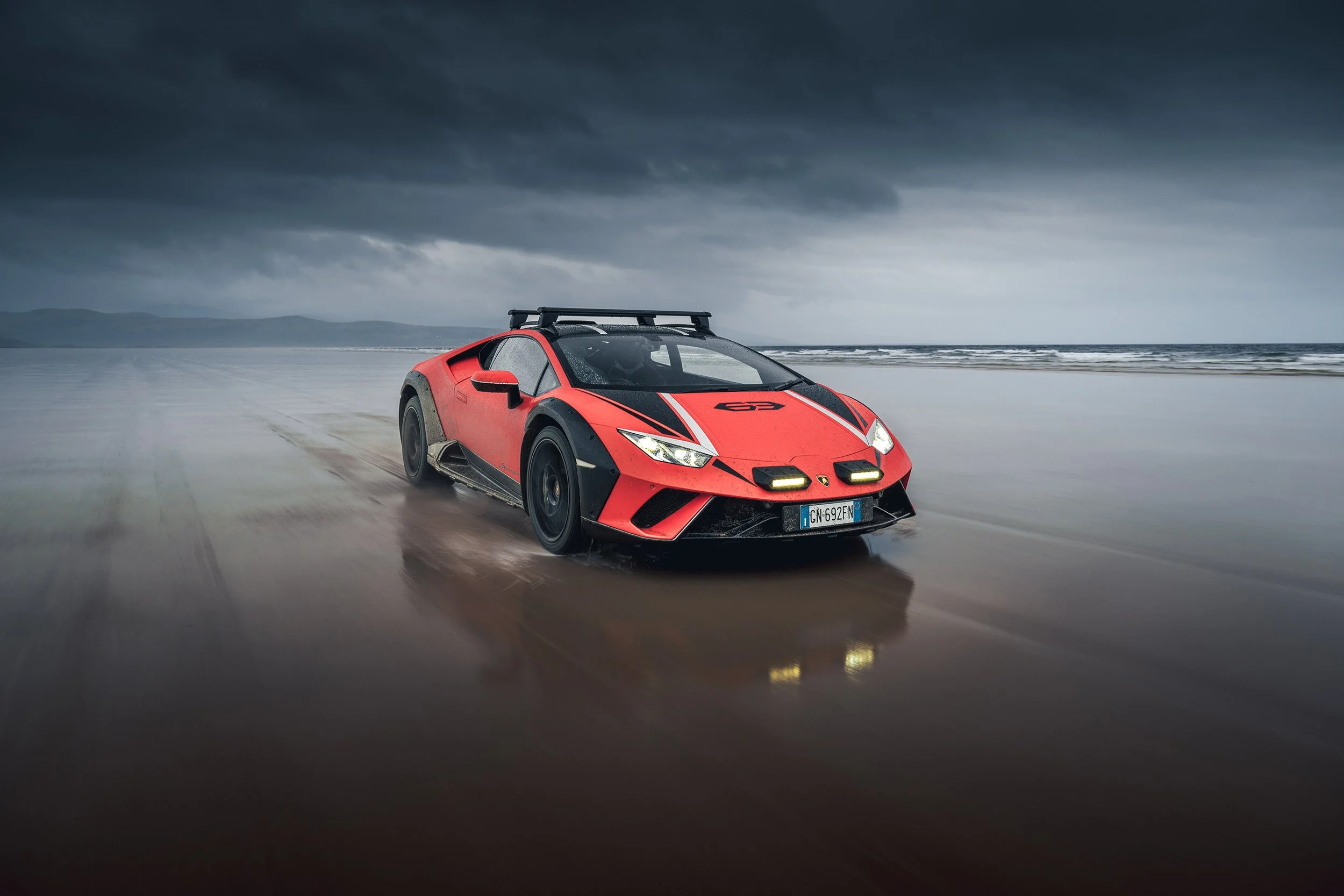 A red and black Lamborghini sports car driving on a wet beach under a dark, cloudy sky.