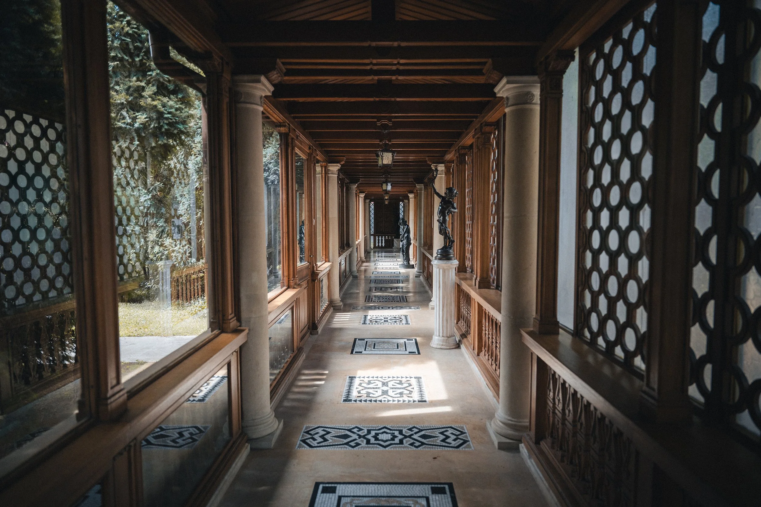 A long, covered corridor with wooden walls, glass windows, classical columns, and decorative statues.