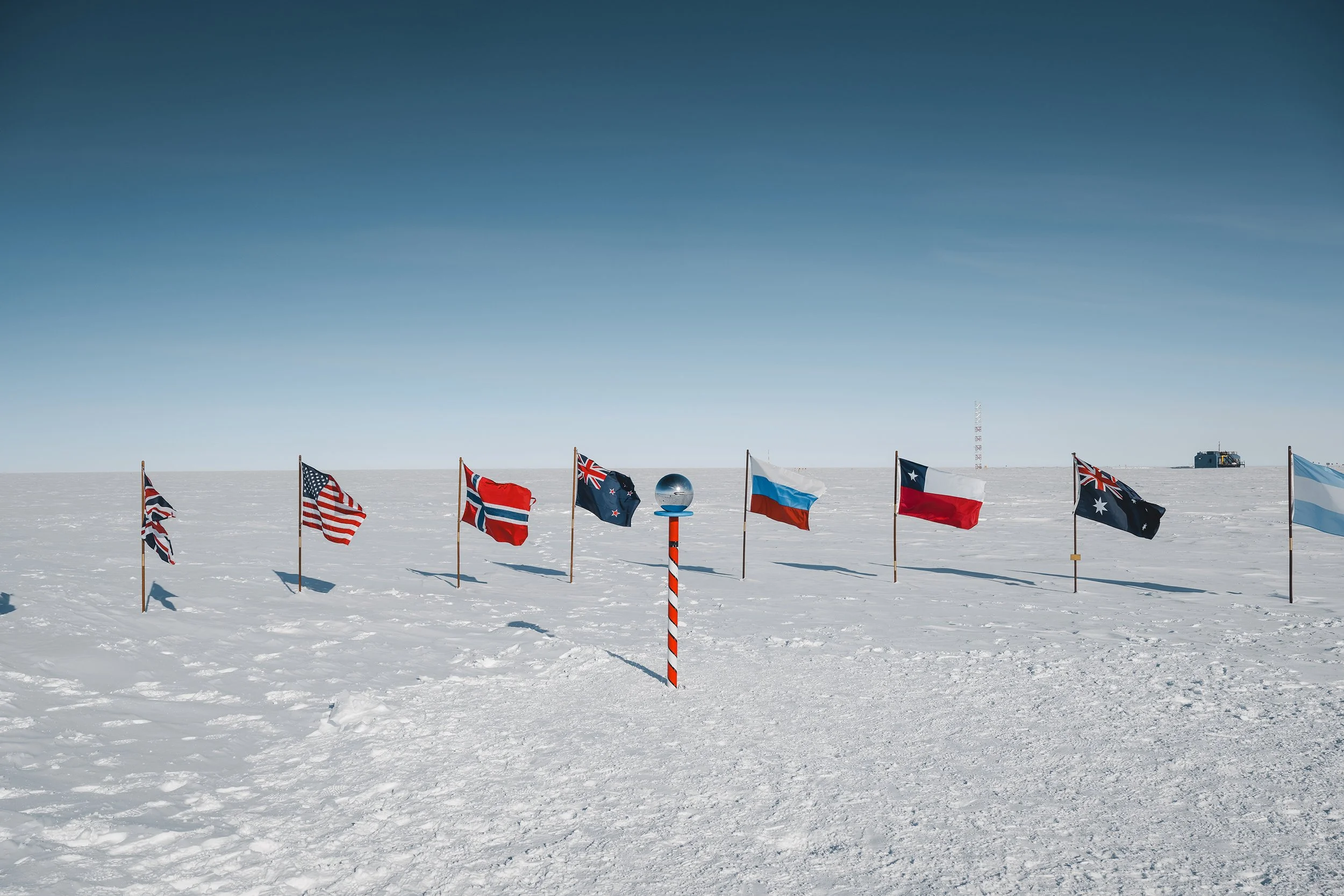 Several international flags standing in the snow on a flat, icy landscape with a clear blue sky. Flags include those of the United Kingdom, United States, Australia, and Russia, among others. A pole with a reflective globe is in the foreground.