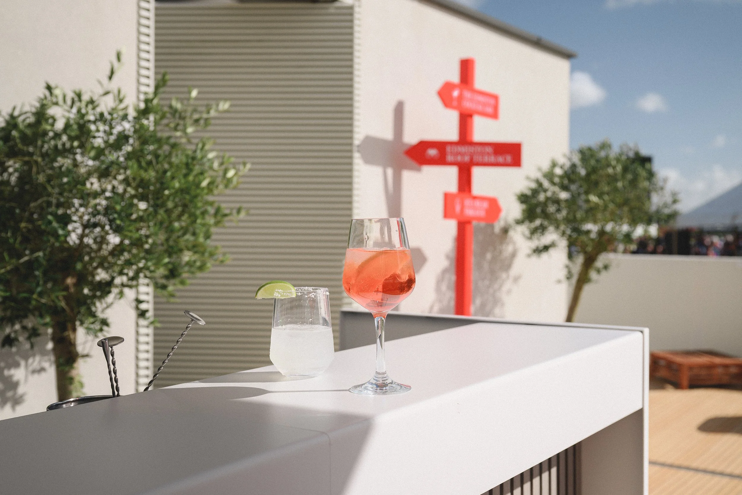 Two cocktails on a white bar counter, with one glass containing a lime wedge and the other a pink drink with a slice of citrus, outdoors on a sunny day with trees and a red signpost in the background.