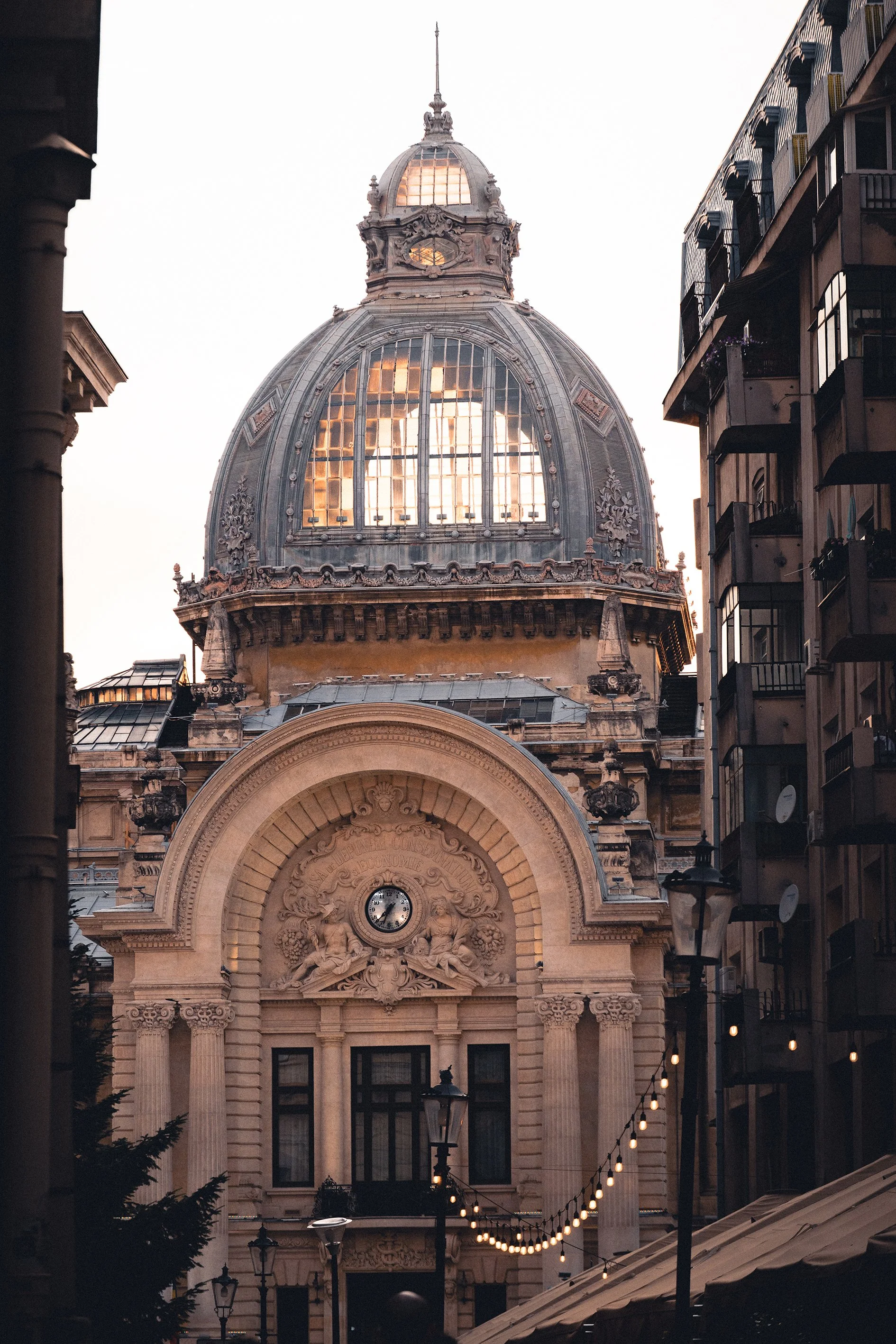 Historical building with ornate stone façade and a large glass dome at sunset, between modern apartment buildings.