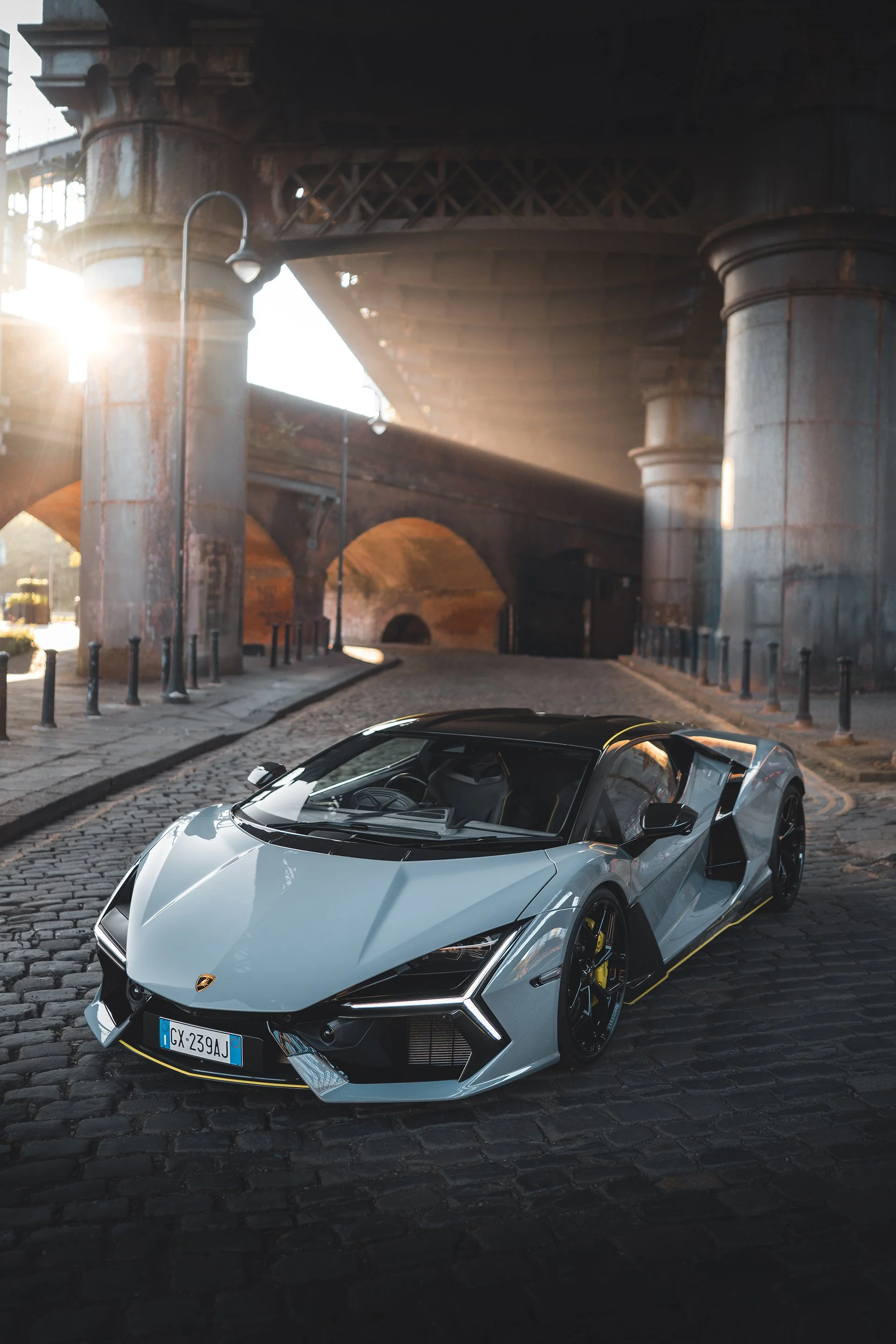 A sleek silver Lamborghini sports car parked on a cobblestone street under an old bridge with large stone pillars, during sunrise or sunset.