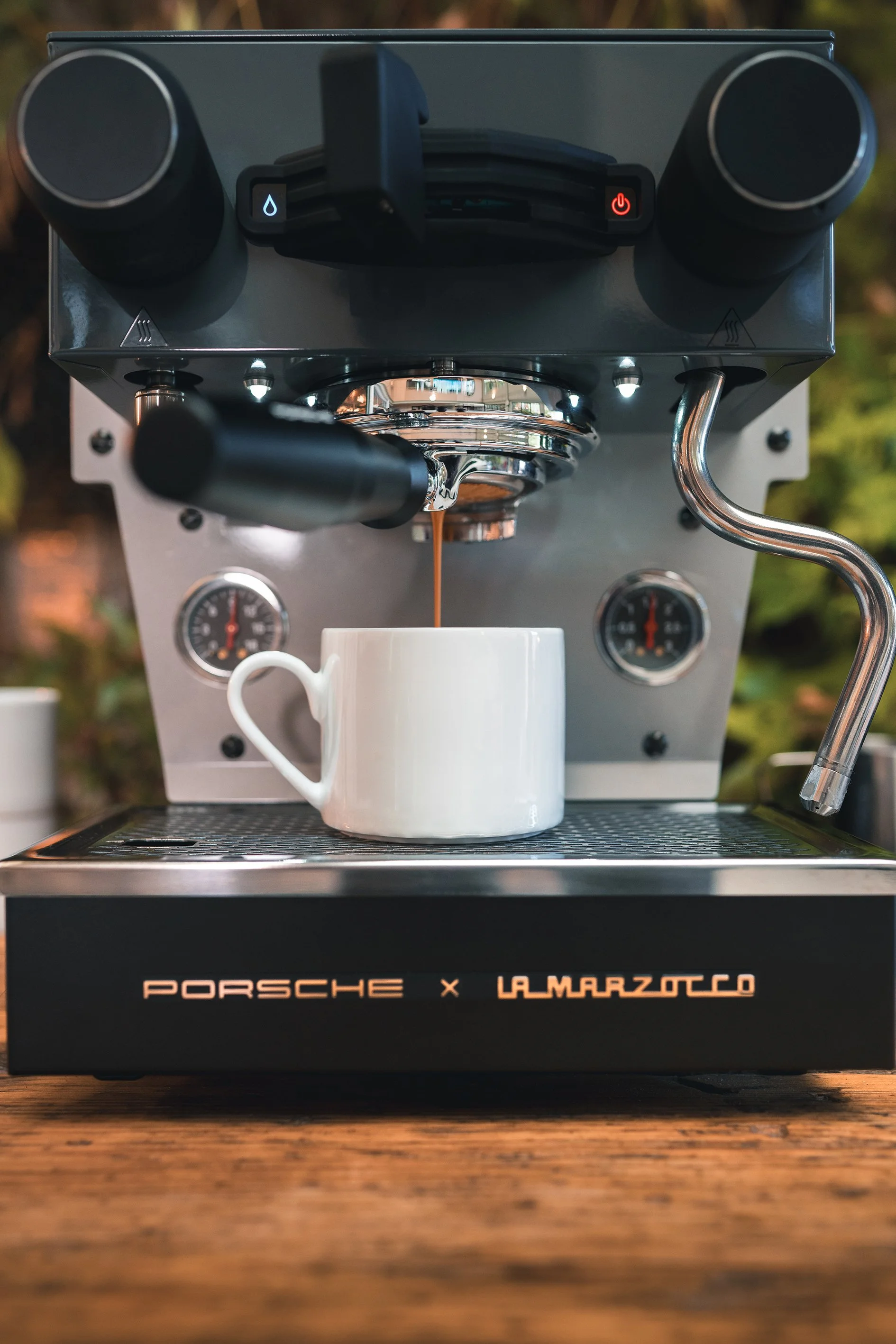 A close-up view of an espresso machine brewing coffee into a white mug, with a logo reading 'Porsche x Lamarzocco' on the front.