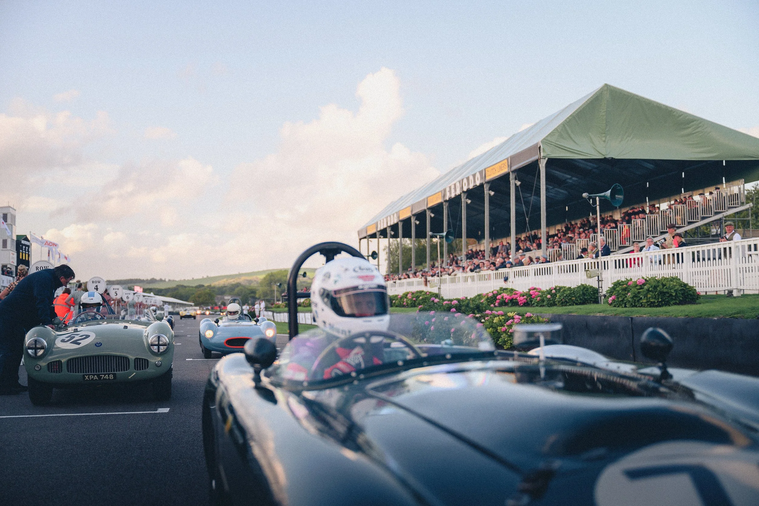 Vintage race cars lined up on a racetrack with a grandstand full of spectators in the background.
