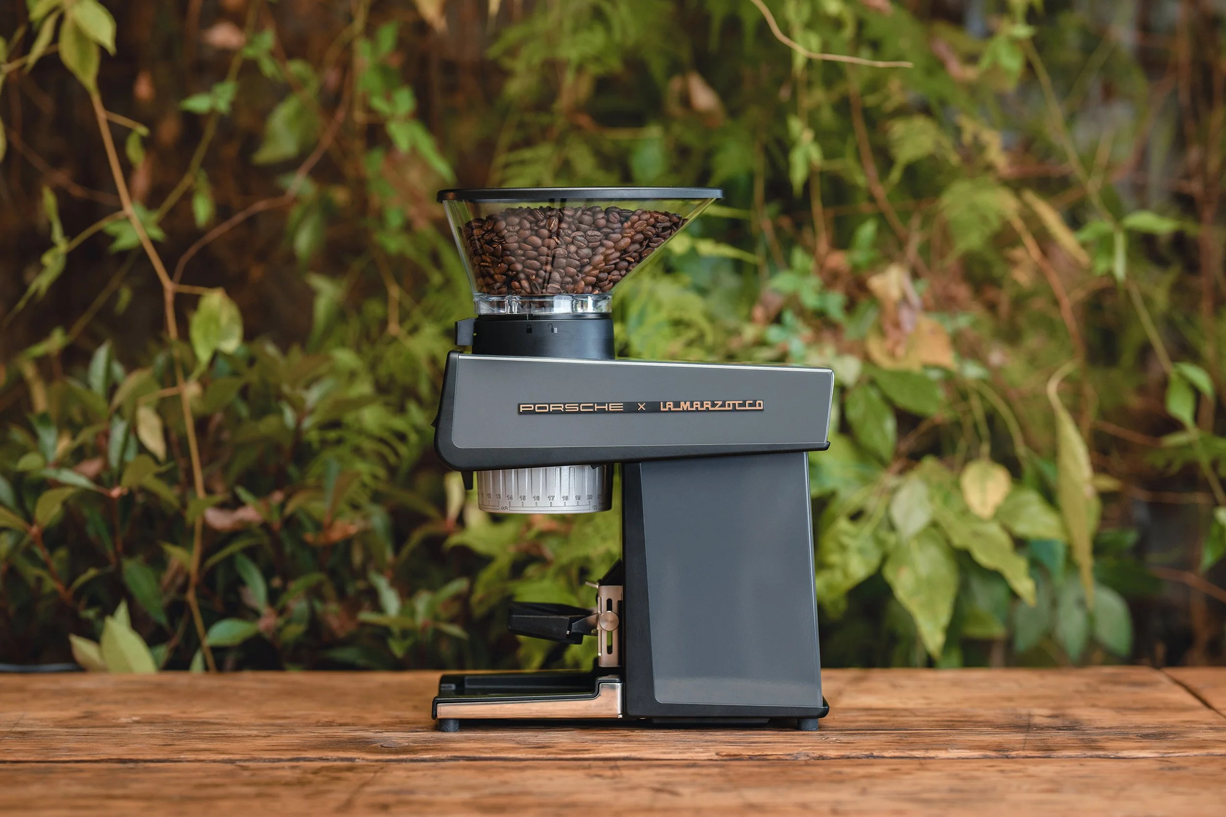 A modern coffee grinder with coffee beans in the top hopper, sitting on a wooden surface with green foliage in the background.