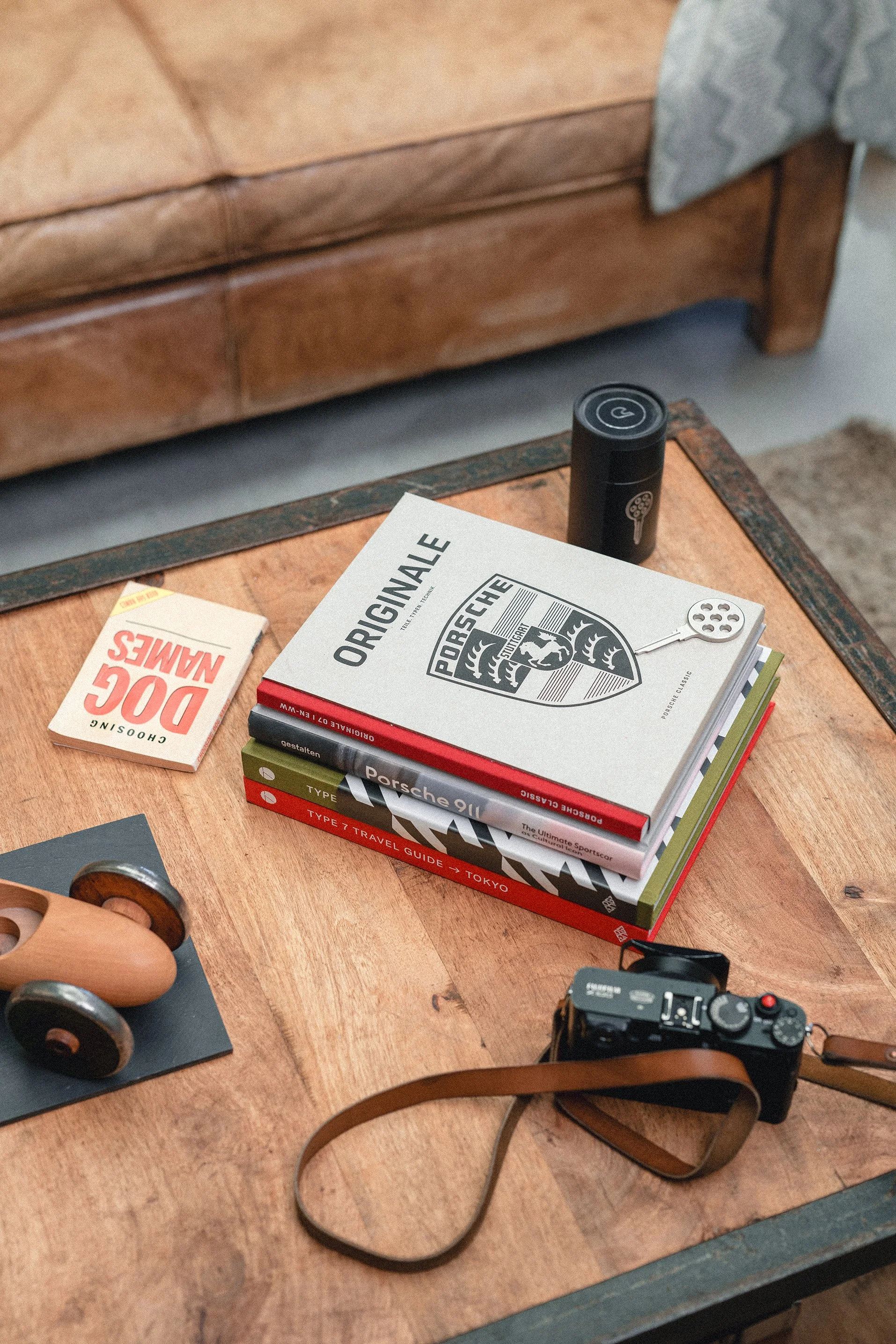 A wooden coffee table with books about Porsche, a camera with a brown strap, a black portable speaker, a toy car, and a pack of chewing gum on it, in front of a tan leather couch.