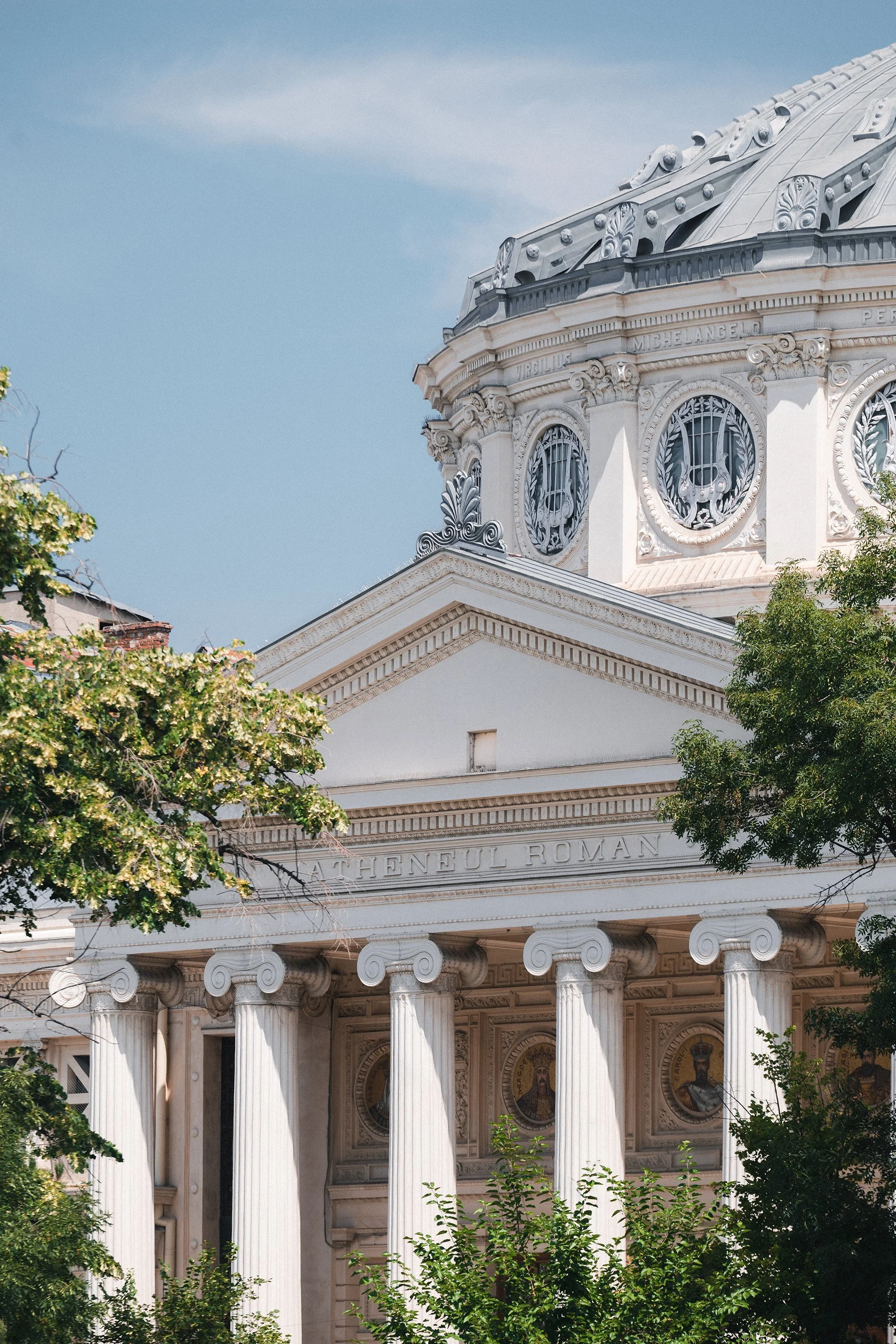 Close-up of a domed, classical-style building with white columns, trees in the foreground, and a blue sky in the background.