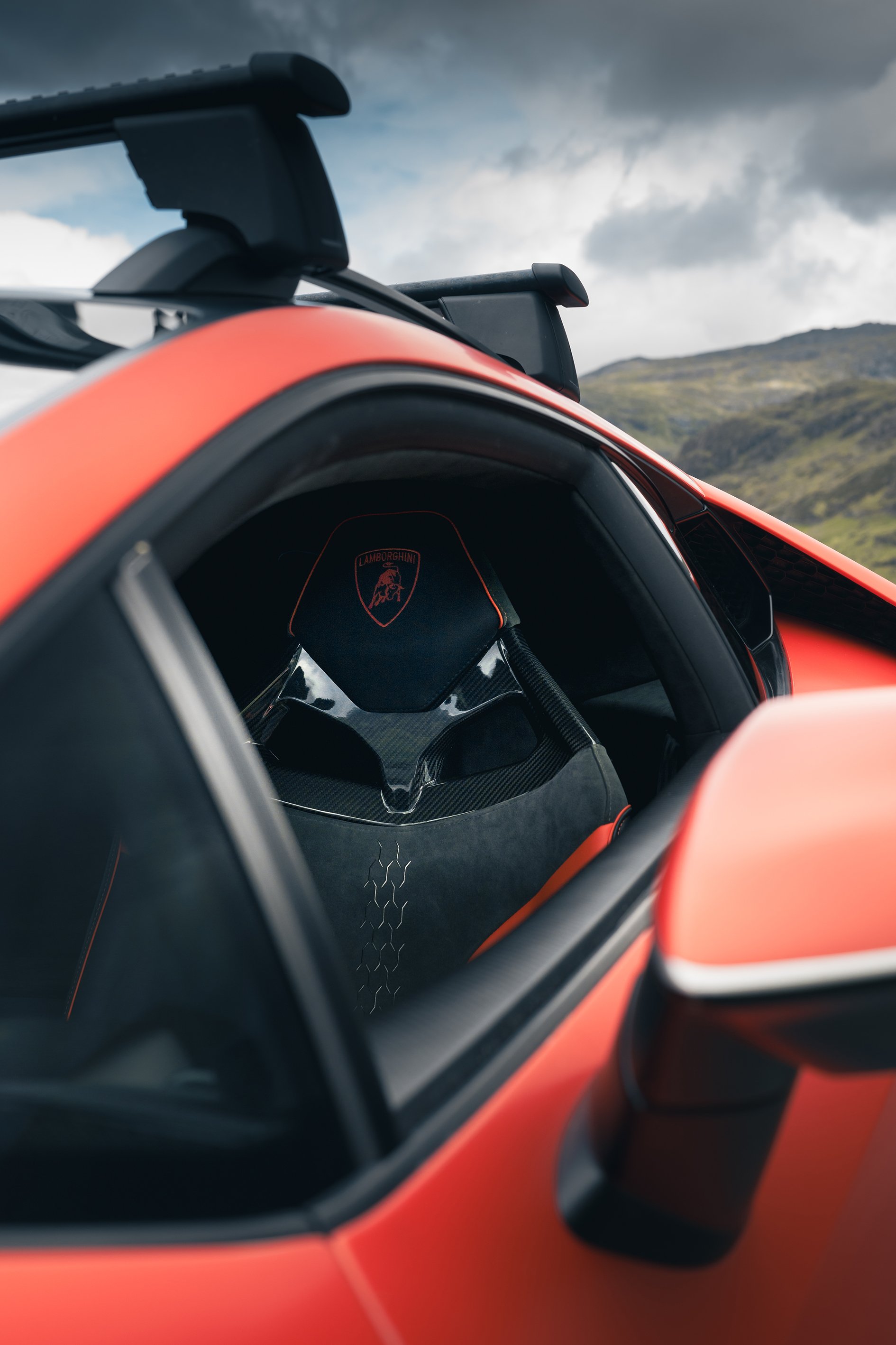 Close-up of a red Lamborghini sports car interior and side mirror with a scenic mountain landscape in the background.