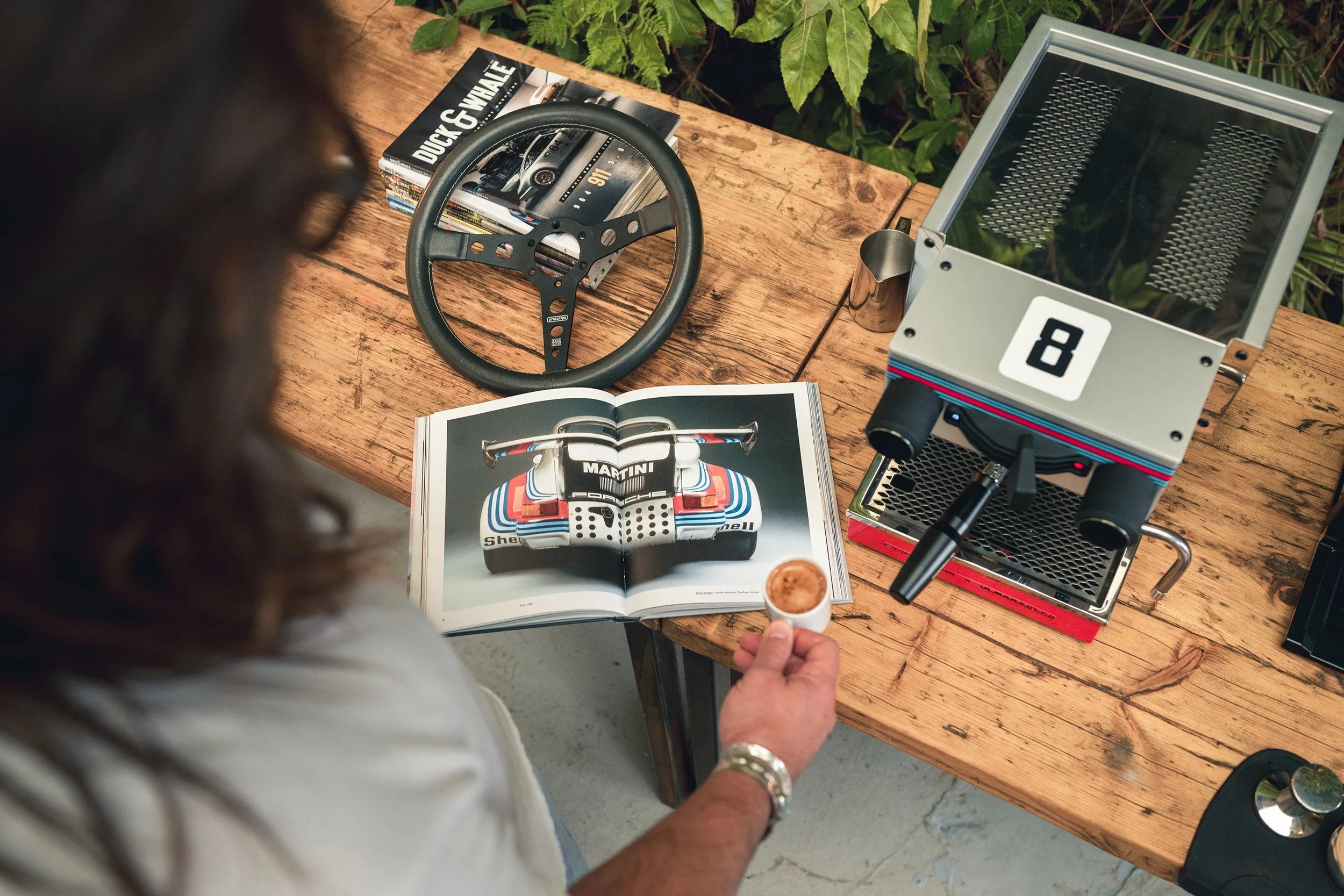 A person holding an espresso cup near a racing book open to a picture of a car's rear wing, set on a wooden table. Also on the table are a racing steering wheel, a gray espresso machine with the number 8, a stack of magazines, and a metal pitcher, wi