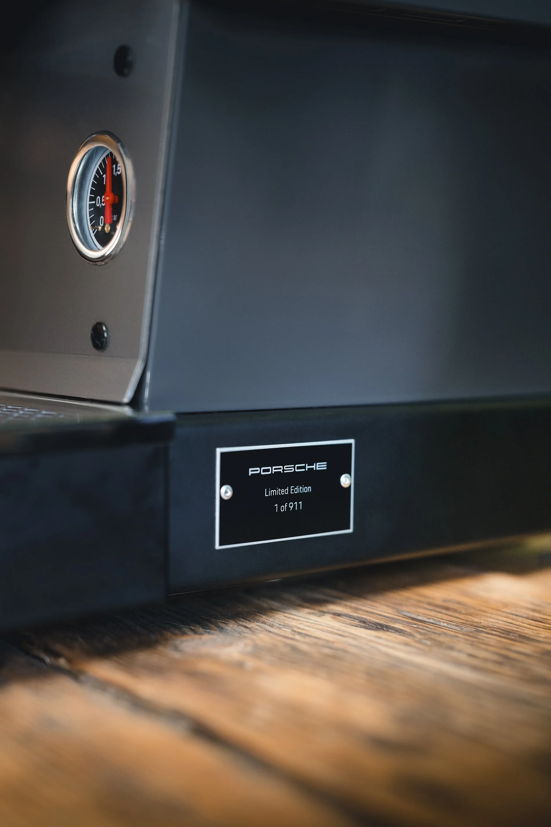 Close-up of a black Porsche limited edition box with a silver plaque indicating 1 of 911, placed on a wooden surface, with a round gauge on the top left.