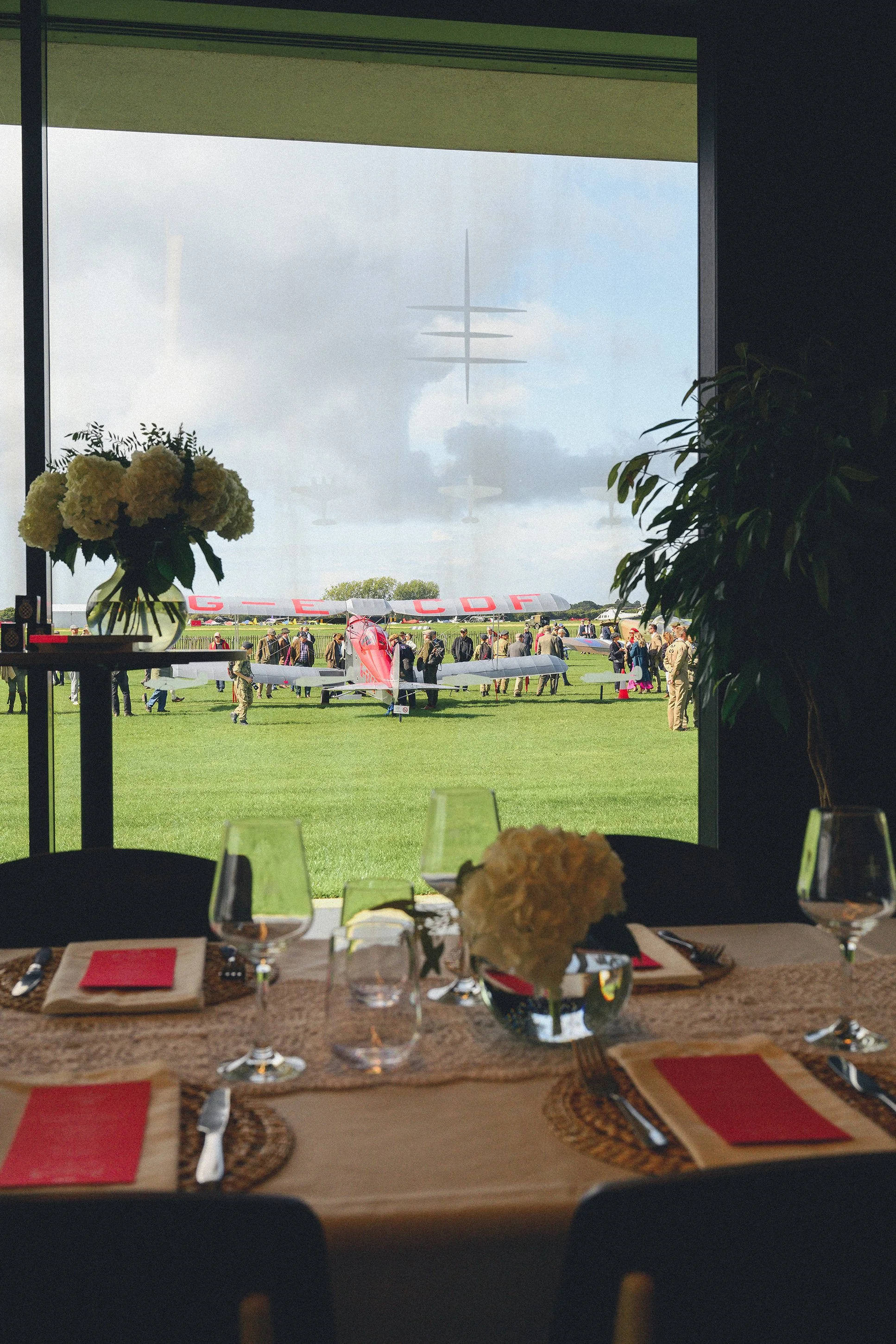 A view of an airplane on the runway seen through a window with a dining table inside the building in the foreground, decorated with flowers, glasses, and place settings. There are crowds of people around the plane, and a red aircraft with white marki