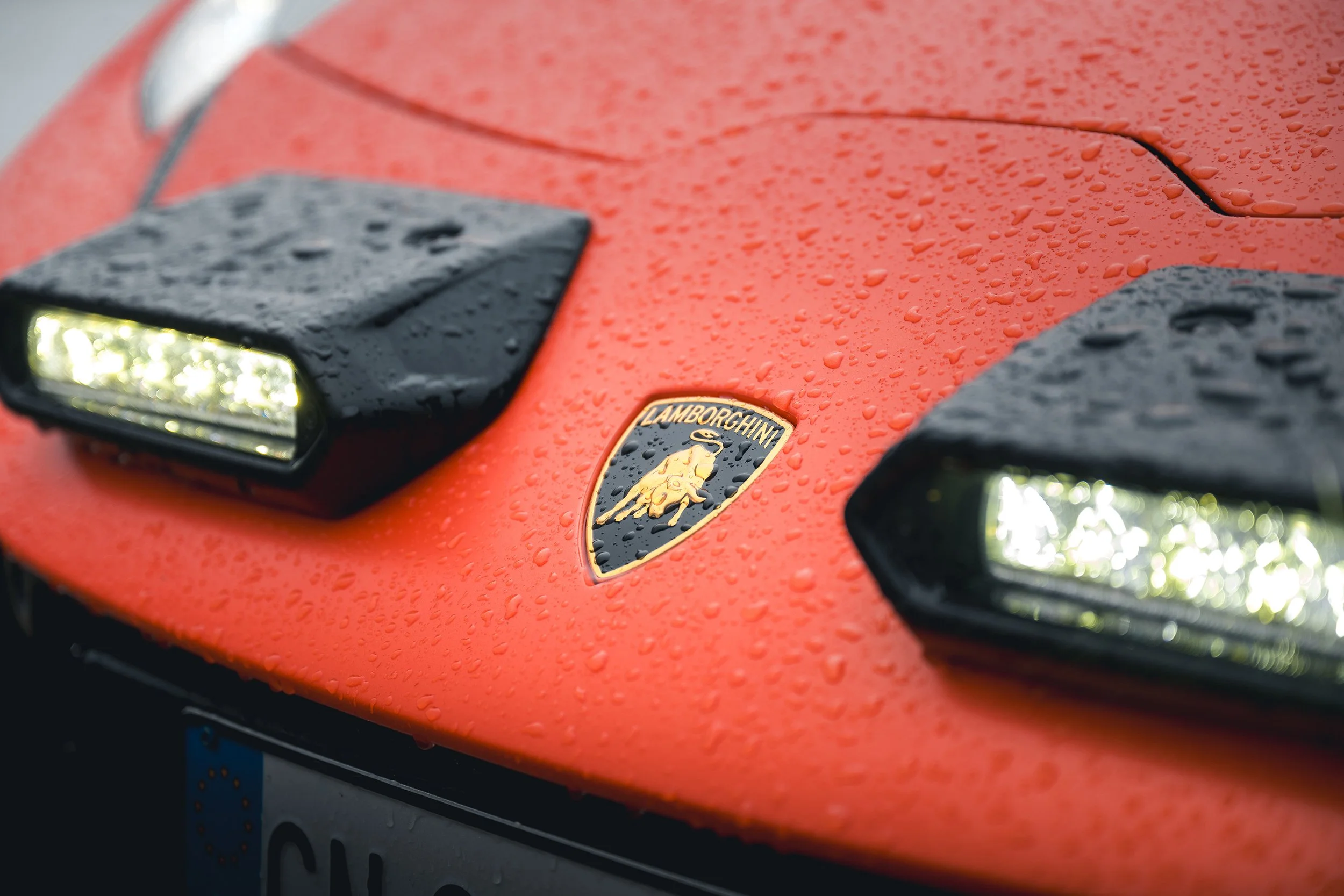 Close-up of a red Lamborghini with raindrops on its surface, featuring the Lamborghini emblem and black aerodynamic vents on the hood.