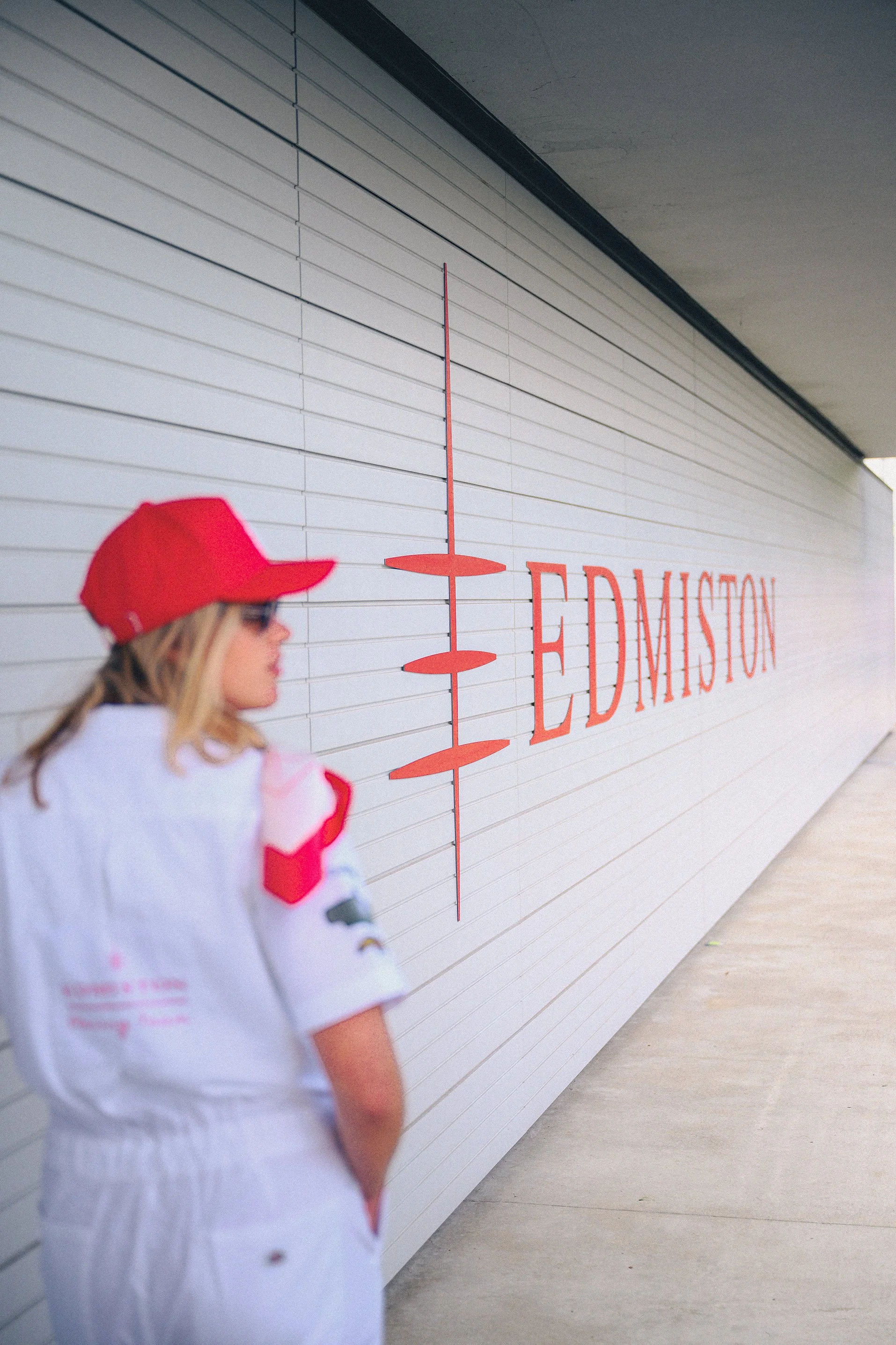 A woman wearing a red cap and white outfit standing in front of a white wall with orange text that says 'EDMISTON' and a logo.