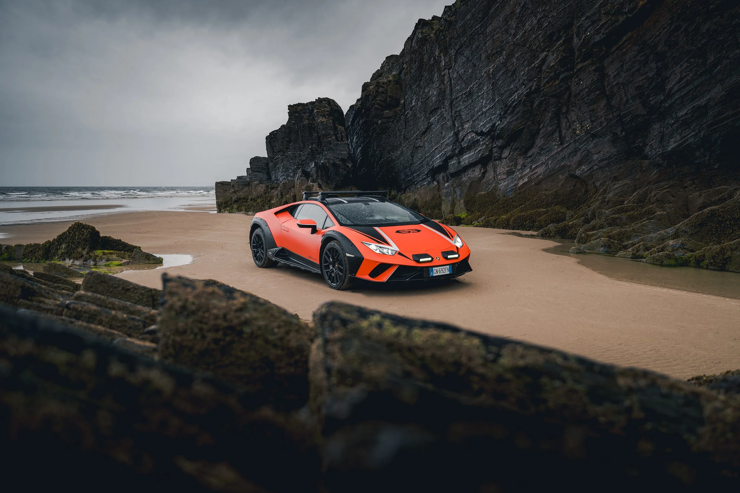 A red sports car parked on a sandy beach near rocky cliffs with an overcast sky.