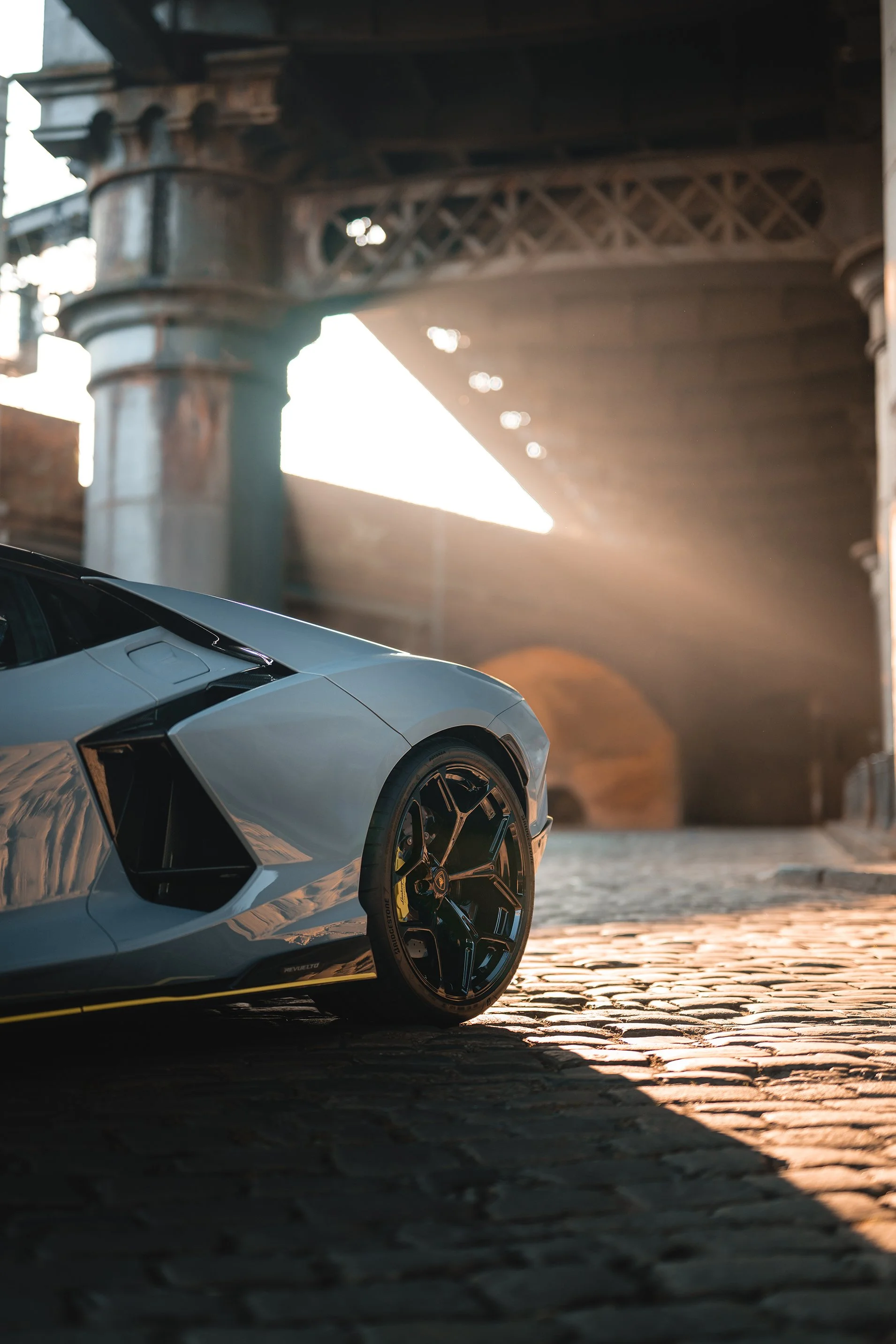 Close-up of a silver Lamborghini sports car parked on a cobblestone street with an urban bridge in the background, illuminated by sunlight.