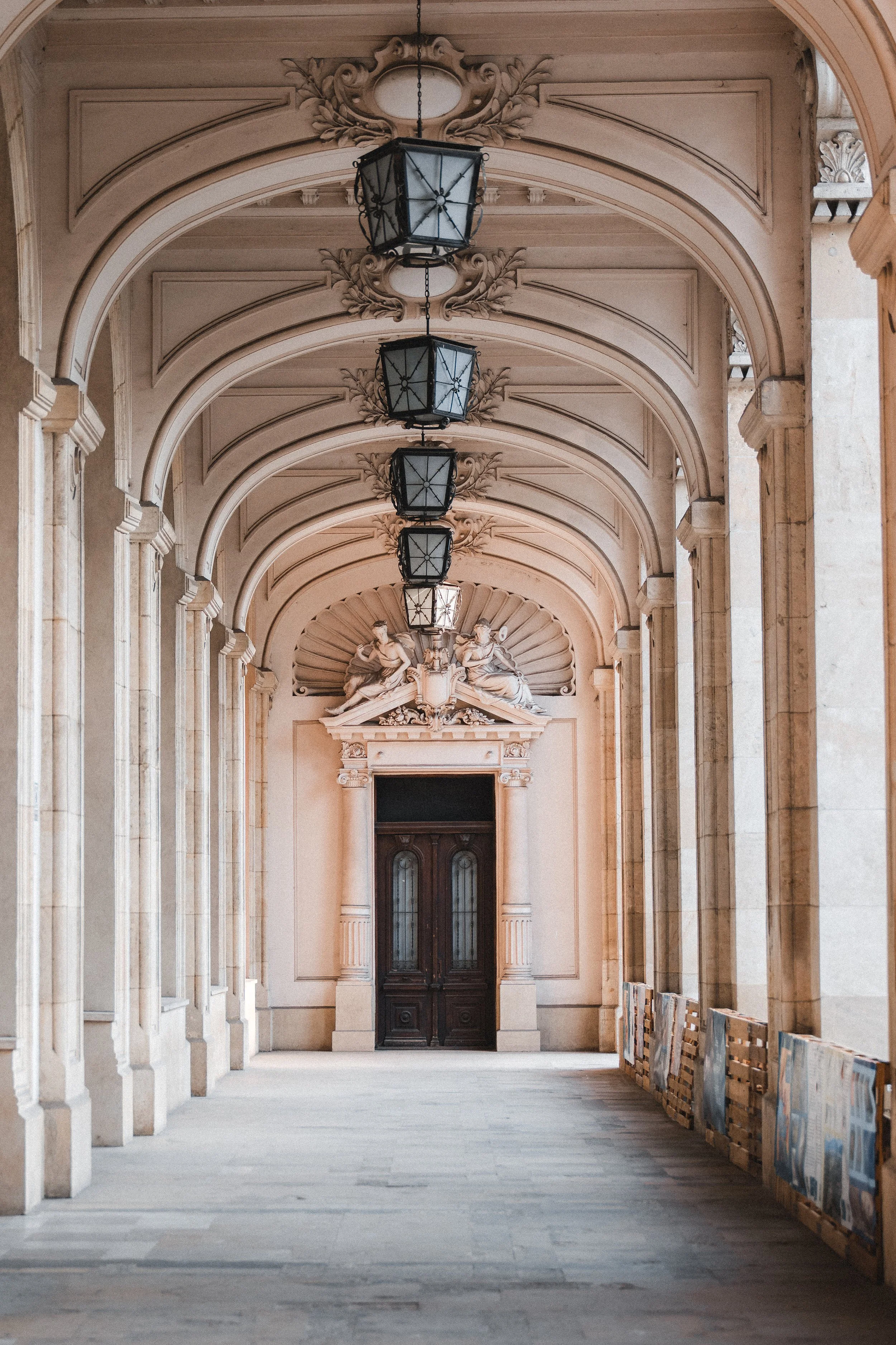 Elegant arched corridor with elaborate ceiling details, hanging black lanterns, and a wooden door framed by classical sculptures and decorative elements.