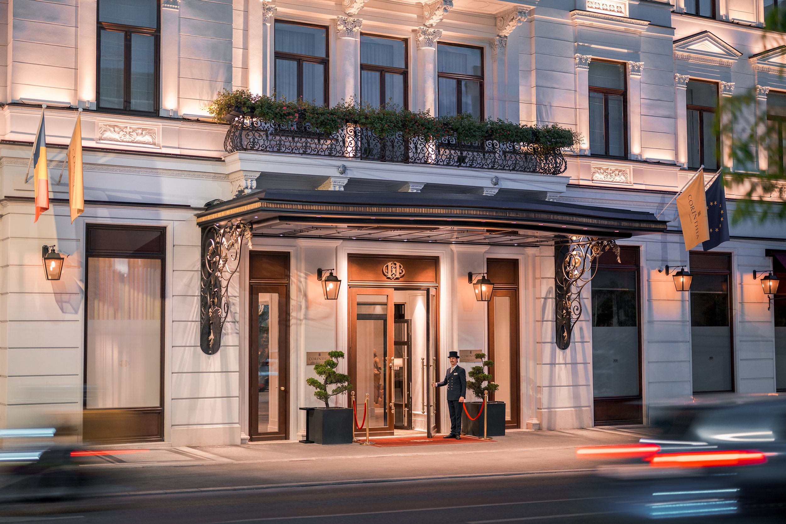 Exterior of an elegant hotel with a white facade, decorative ironwork, small trees at the entrance, and a doorman in a uniform. The building has large windows, flower boxes, and flags on the sides.