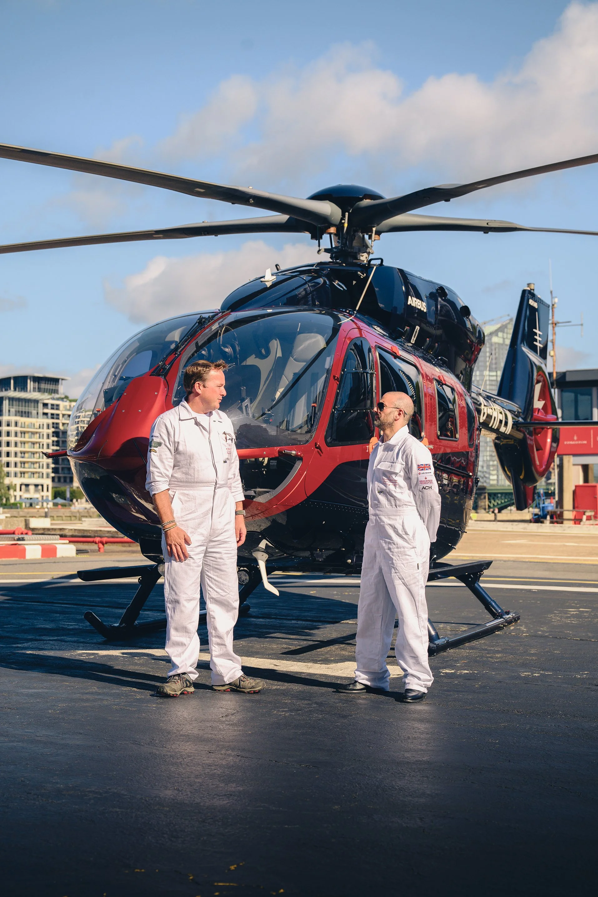 Two men in white pilot suits standing on a helipad beside a red and black helicopter, with a cityscape in the background under a partly cloudy sky.